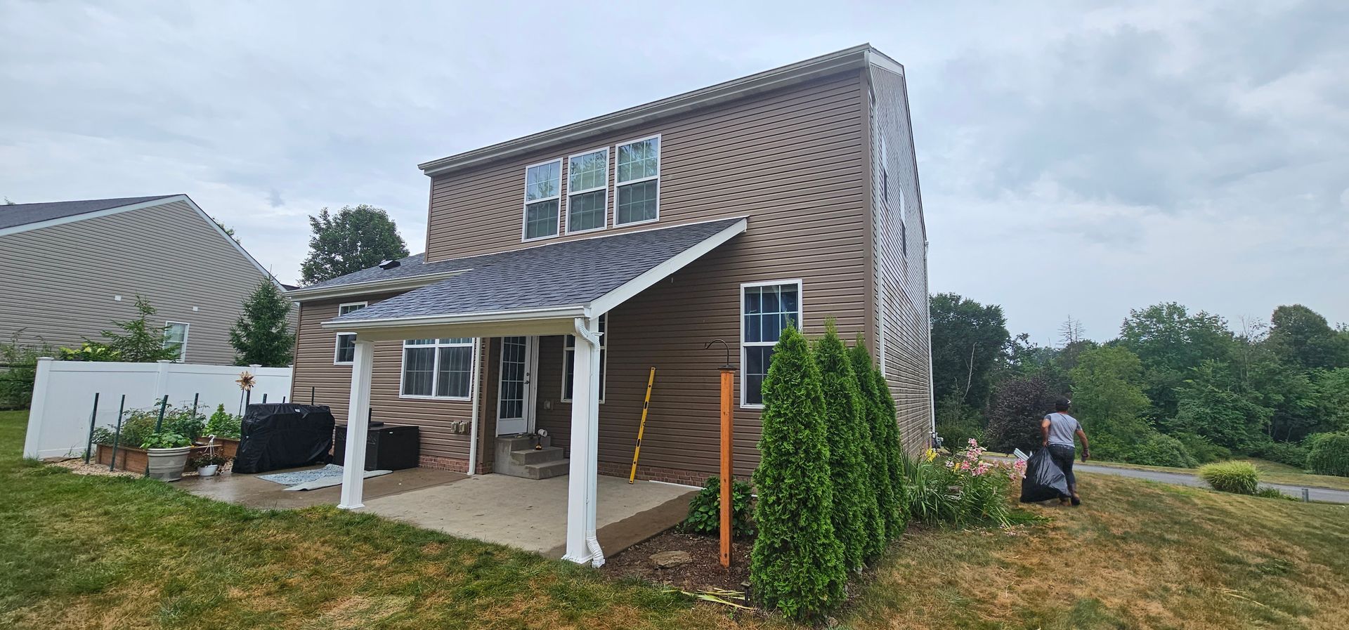 Two-story house with brown siding and a porch. A person bags yard waste in the background. Overcast sky.