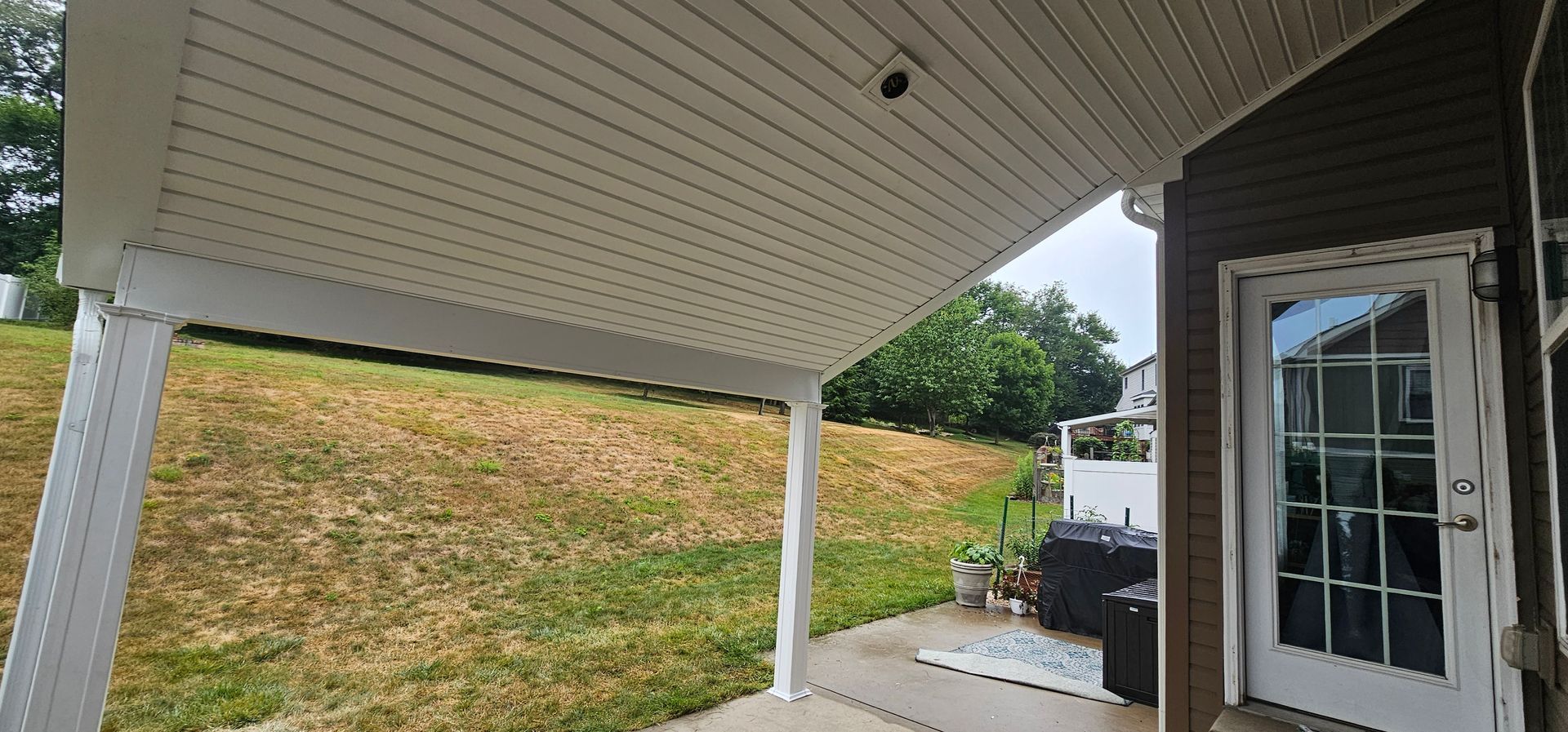 View from under a covered patio with a white ceiling and support posts, overlooking a grassy yard.