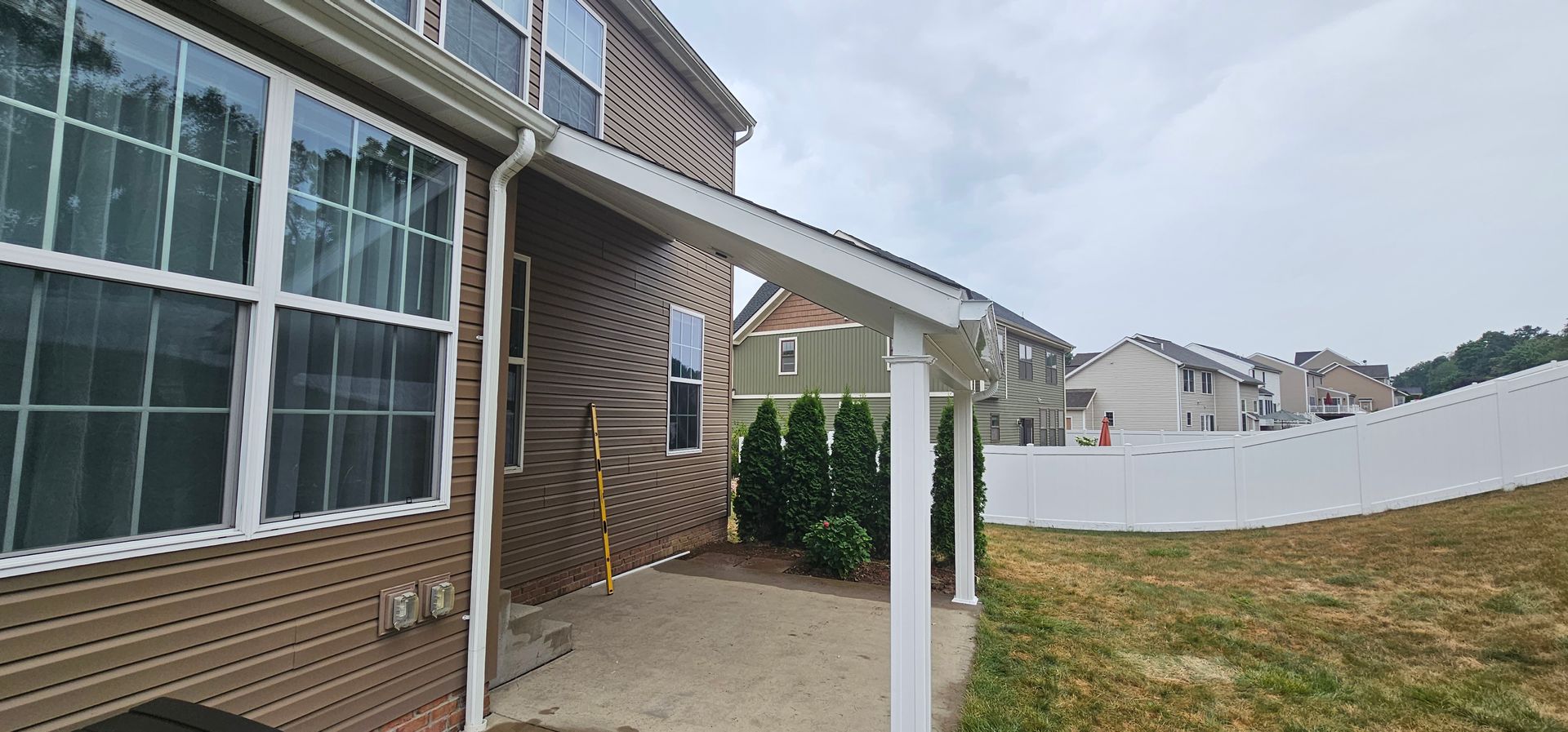 Backyard view of a house with a covered patio, green bushes, and a white fence under an overcast sky.