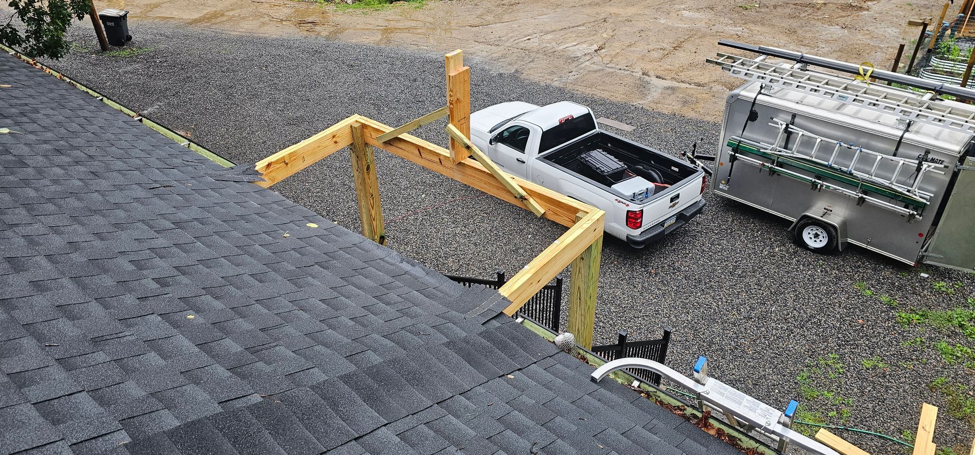 Wooden structure extending from a roof, a white pickup truck, and a trailer on gravel.