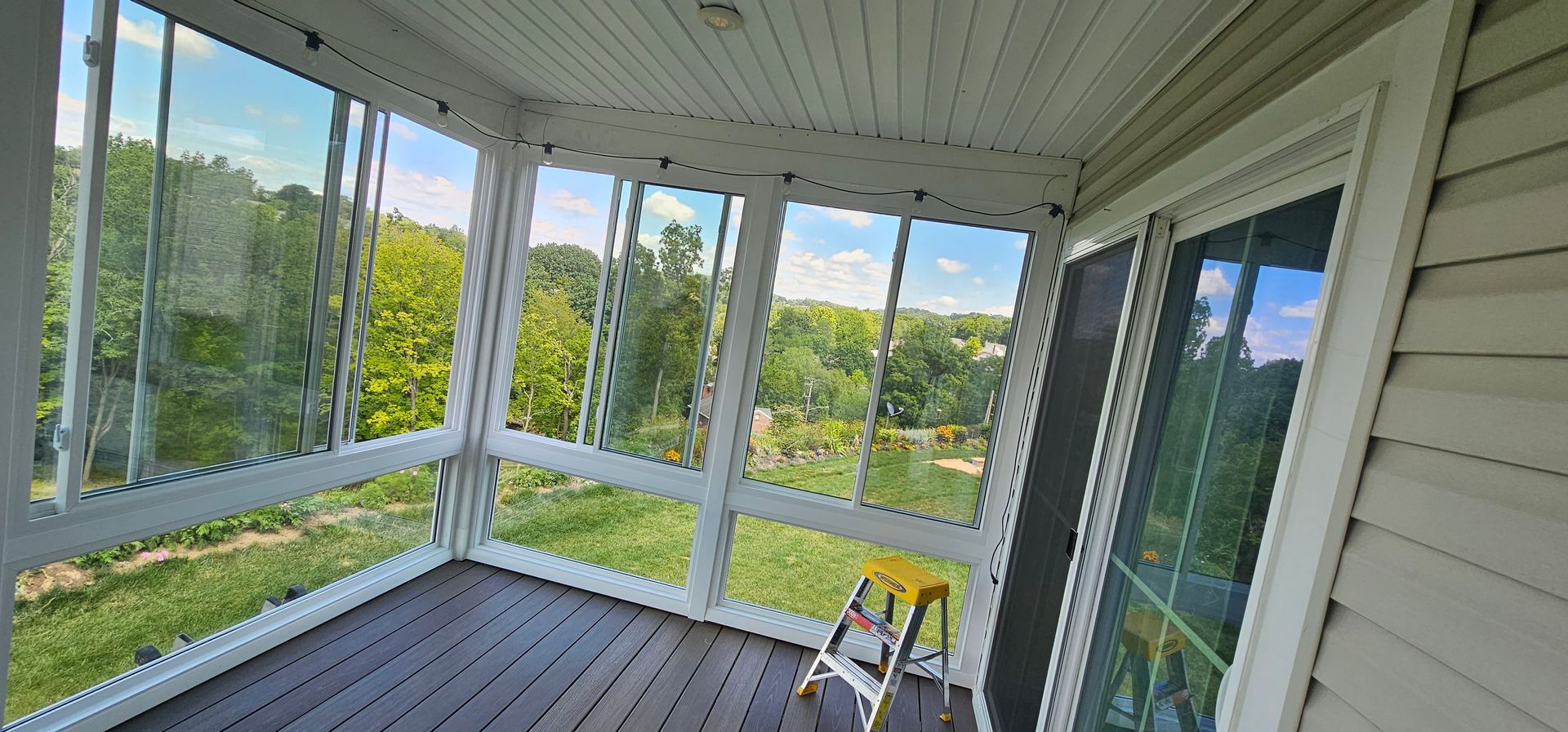 Screened-in porch with windows overlooking a green landscape and blue sky. A yellow ladder sits inside.