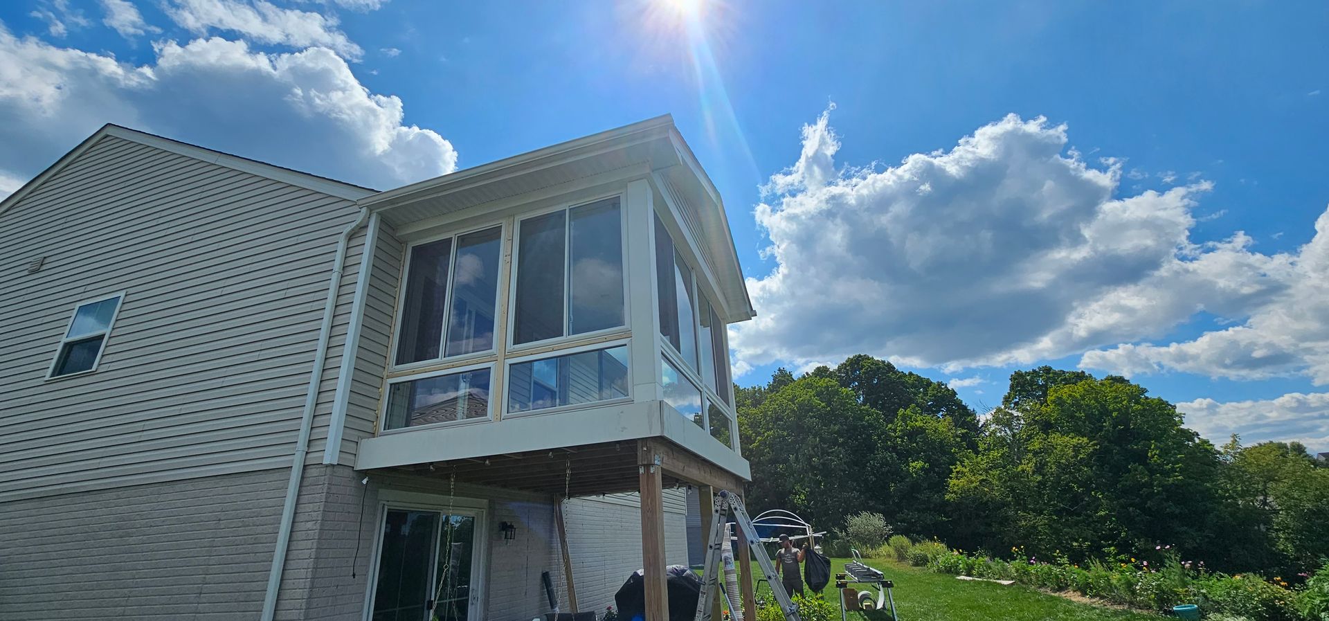 Screened-in porch on a house with blue sky and bright sunlight. Trees and a garden are visible.