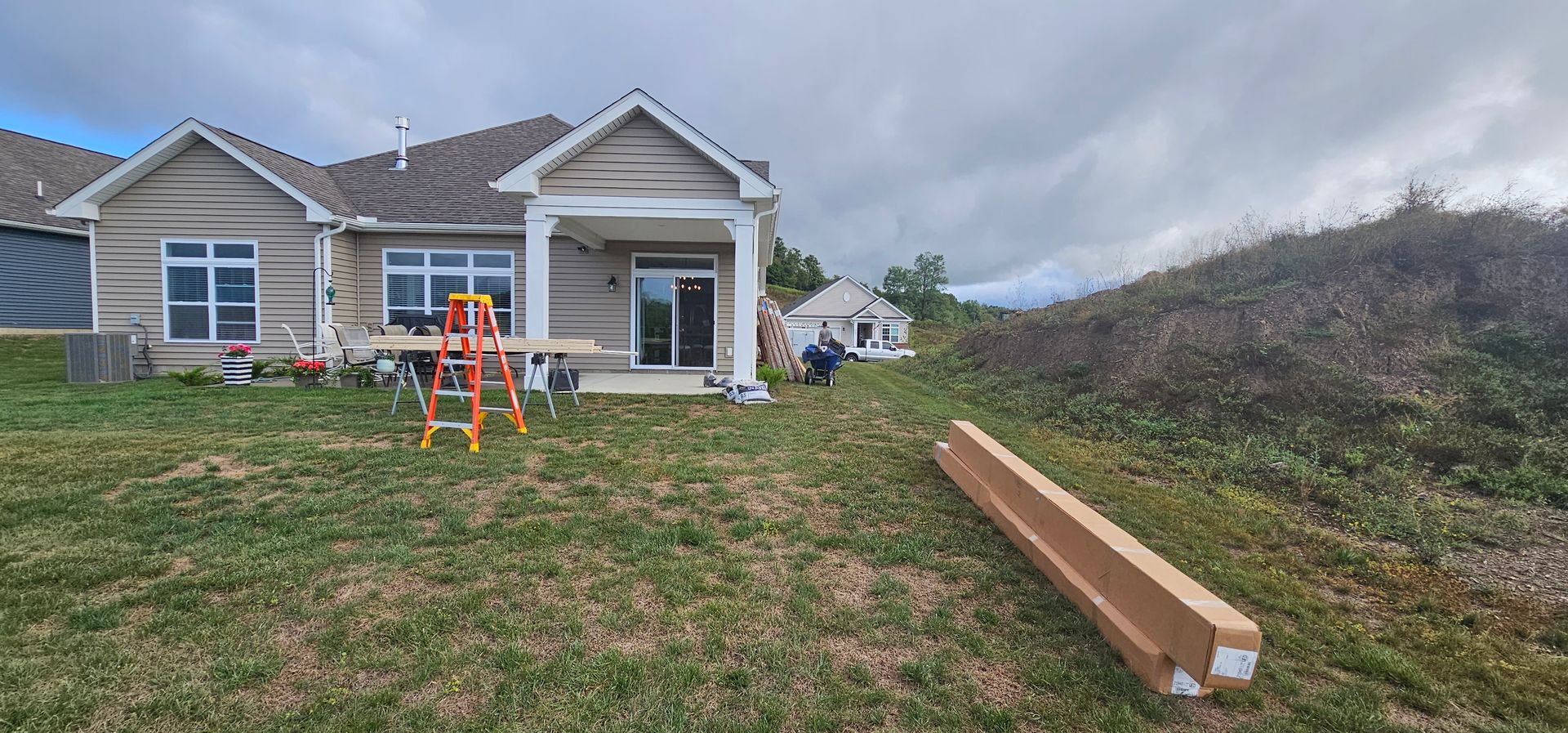 Backyard with house and construction. Brown cardboard, orange ladder on the grass. Overcast sky.