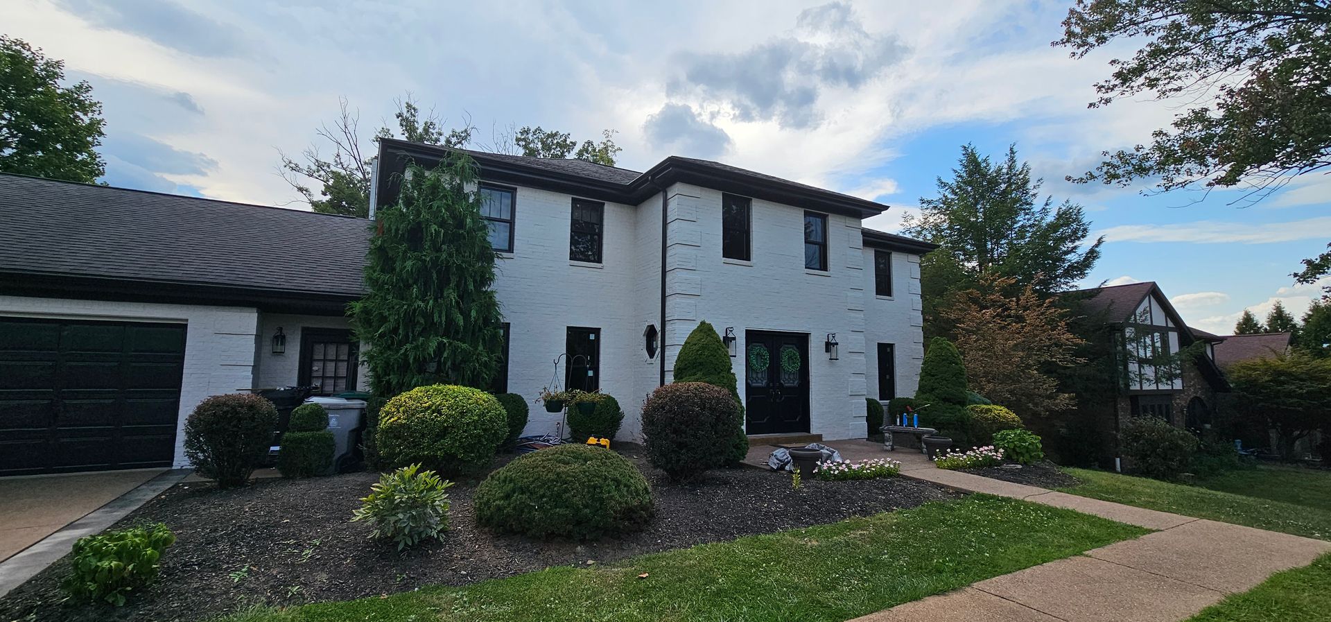 Two-story white brick house with dark roof and trim, black front door, lush landscaping, and green lawn.