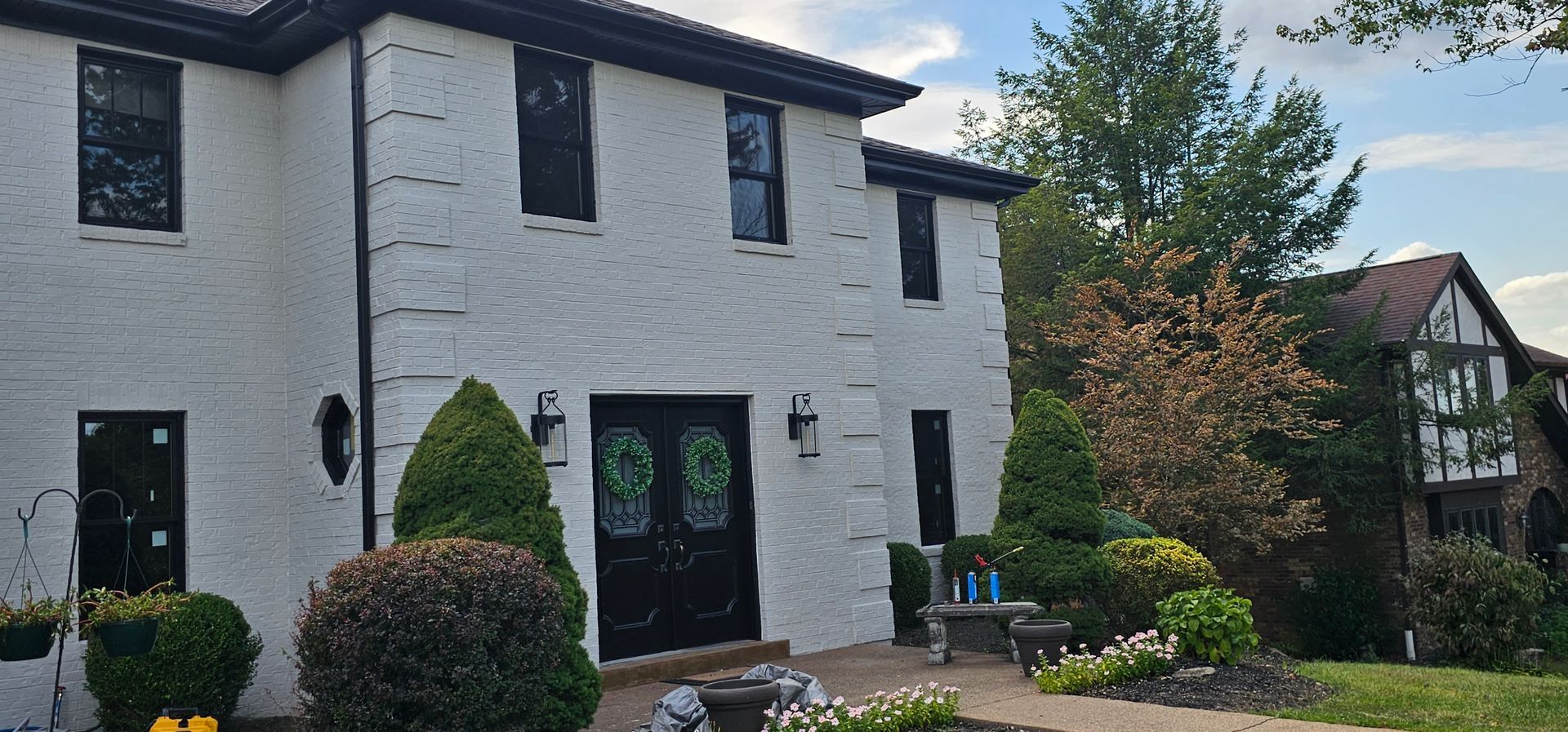 White brick house with black trim and door, two stories, front yard with trees and another house on the right.