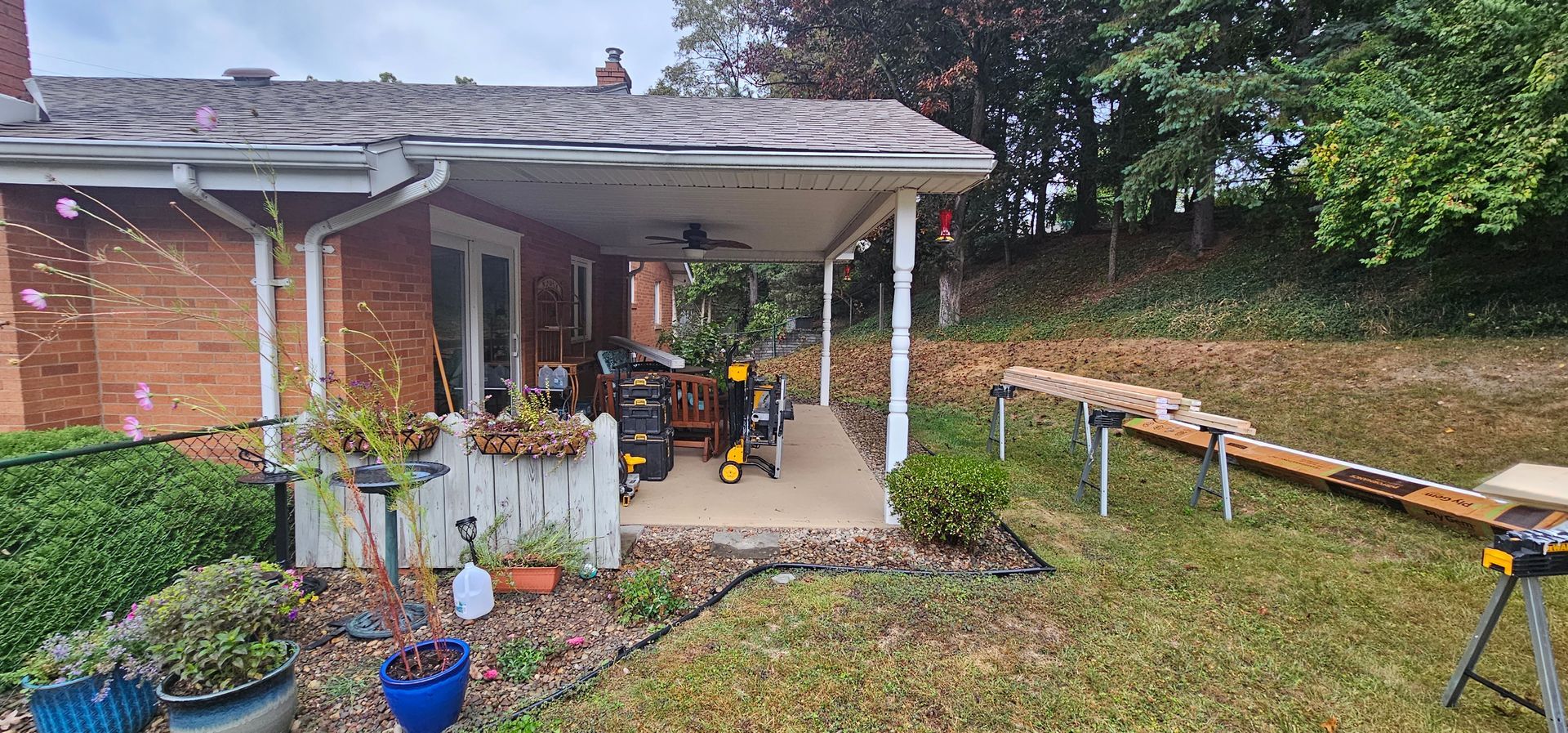 A brick house with a covered patio, tools, and a wooden plank extending onto a lawn with trees.
