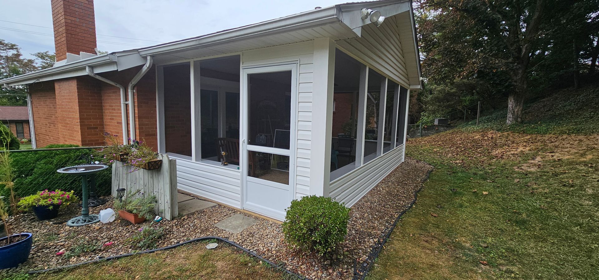 Screened-in porch attached to a brick house with a grassy yard and trees in the background.