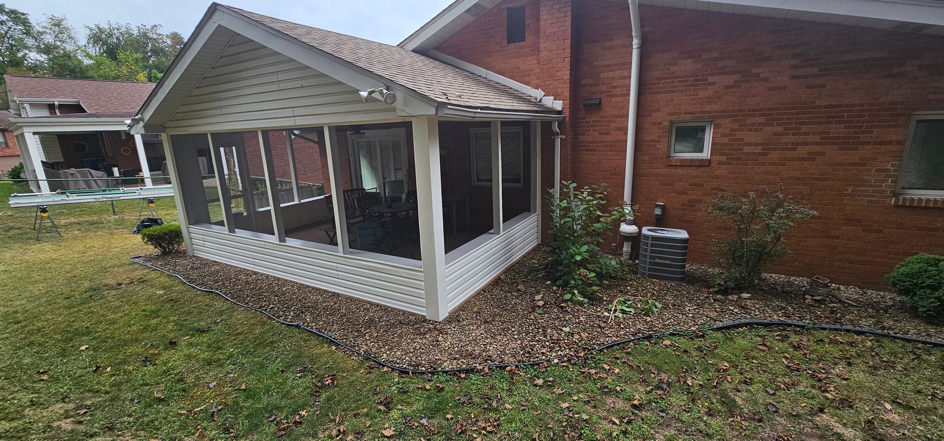 Screened porch attached to a brick house. Landscaping with stones borders the porch. Green grass and trees are in the background.