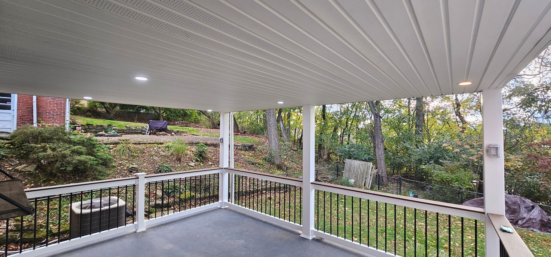 Covered porch overlooking a wooded area. Black railing, white columns, and ceiling with recessed lights.