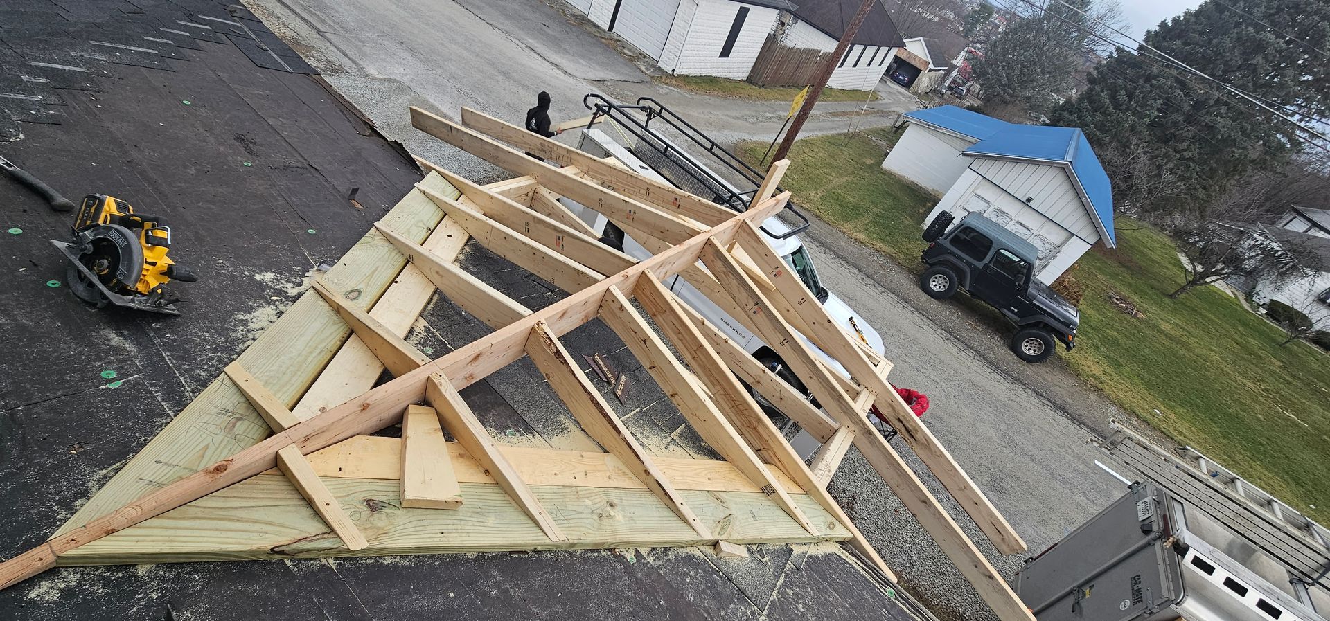 Roof under construction with wooden framing and a circular saw, black vehicle, and small blue building.