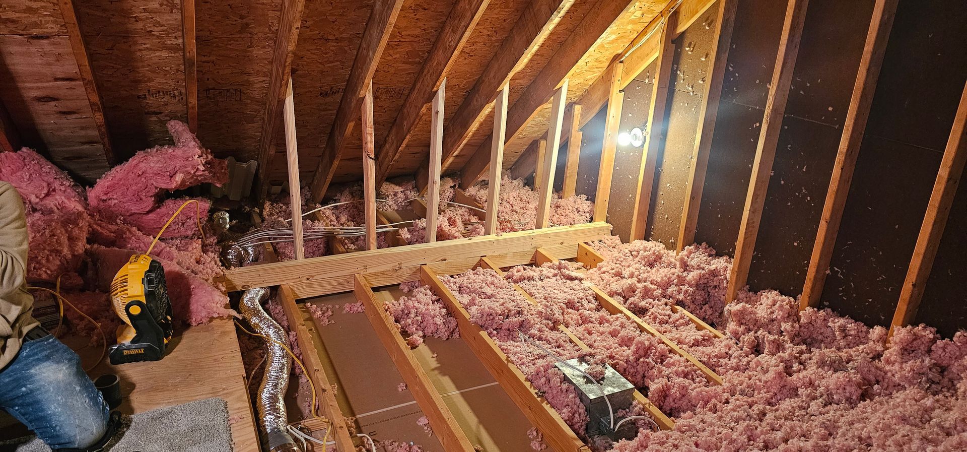 Attic filled with pink insulation, wooden beams and rafters. A work space with a saw and other items.