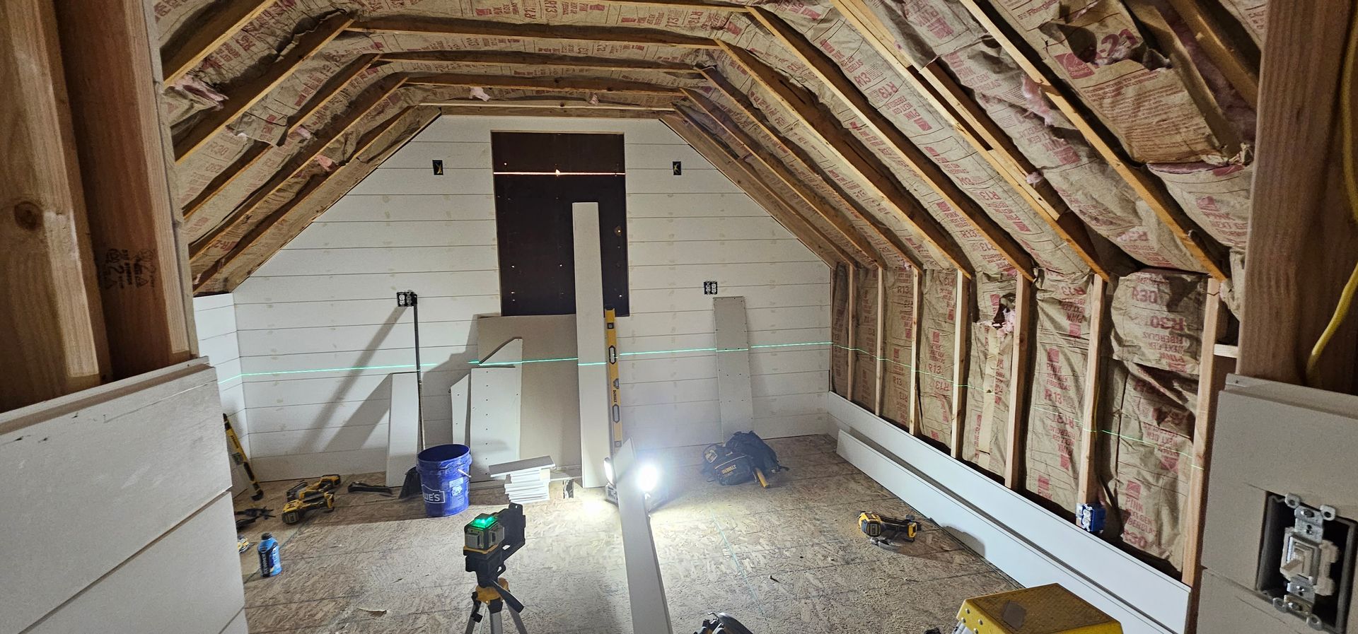 Interior of a room under construction with exposed wooden beams, insulation, and drywall. A laser level is set up.