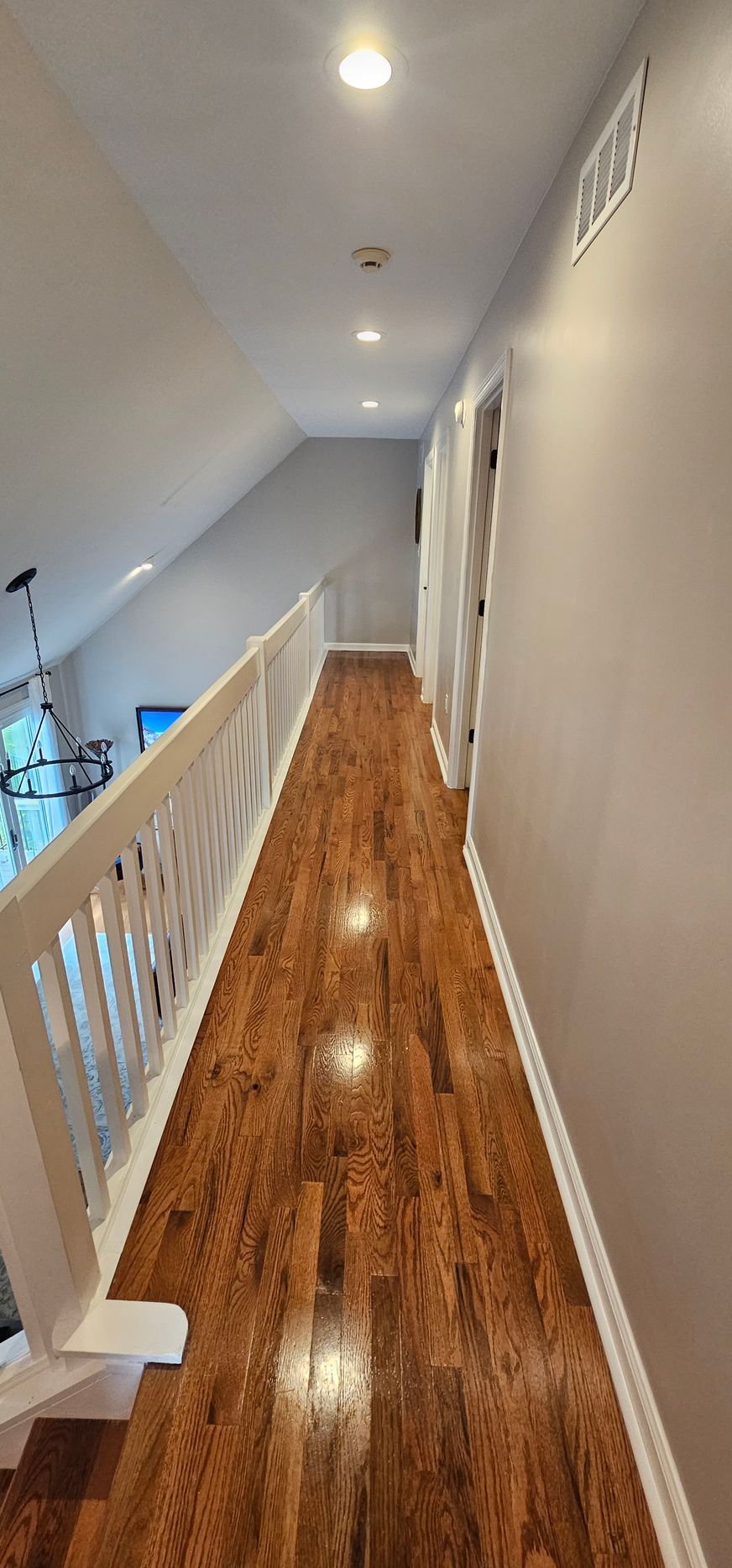 Narrow hallway with wood floors, white railing, and recessed ceiling lights. Gray walls and a door are visible.