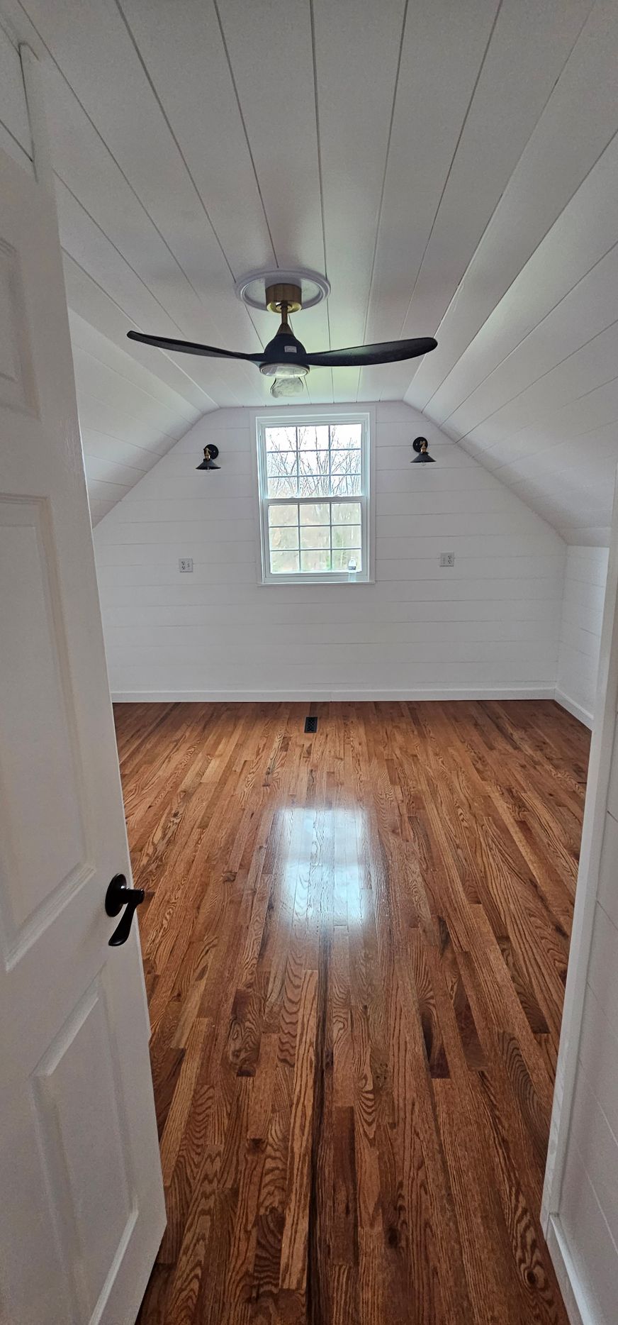 A small room with wooden floors, a ceiling fan, and a window. White walls and door frame.