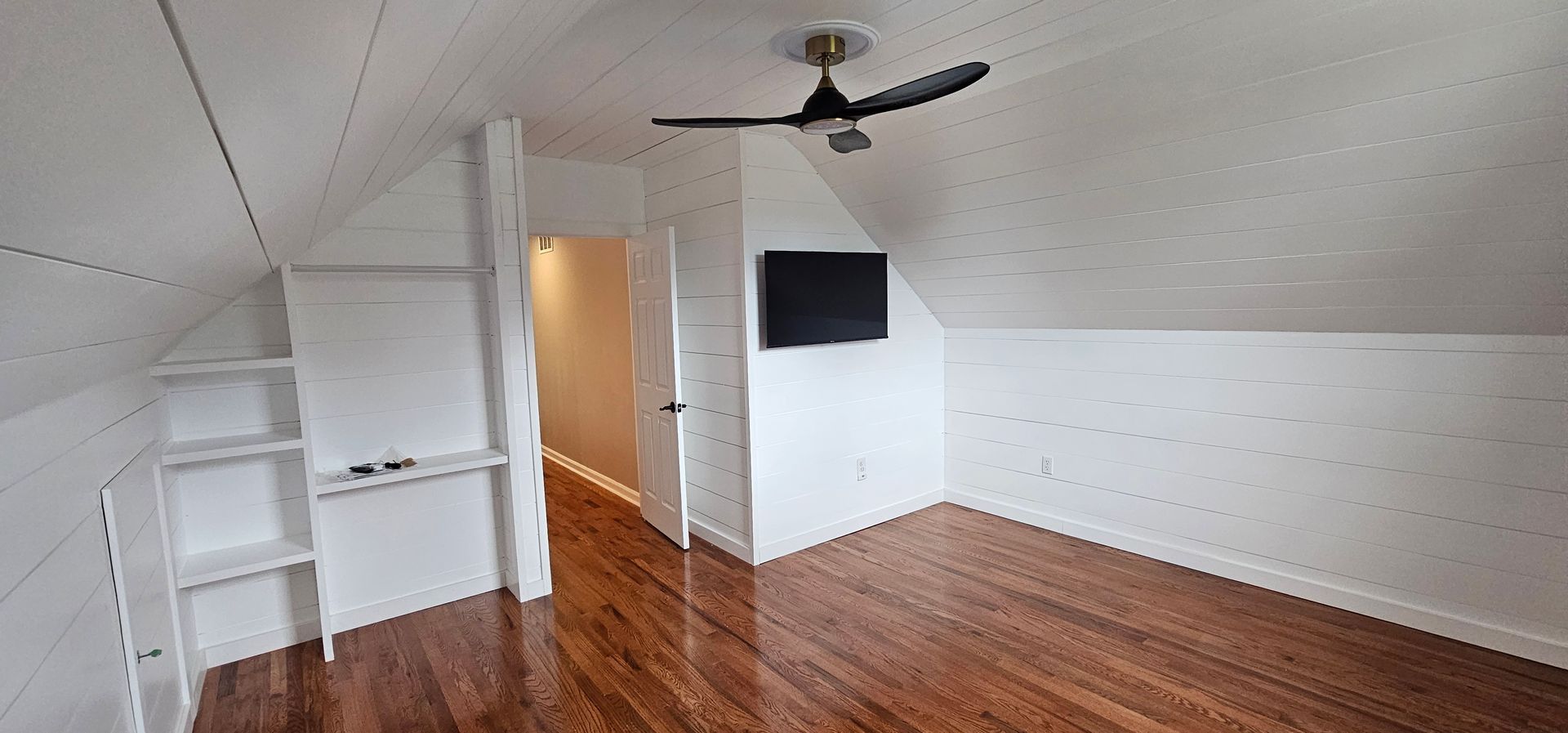 Bedroom with sloped ceiling, wooden floors, built-in shelves, and a wall-mounted TV. A ceiling fan is visible.