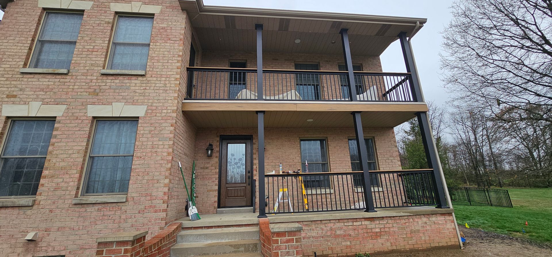 Two-story brick house with a front porch and balcony. Brown door and black railings. Overcast day.