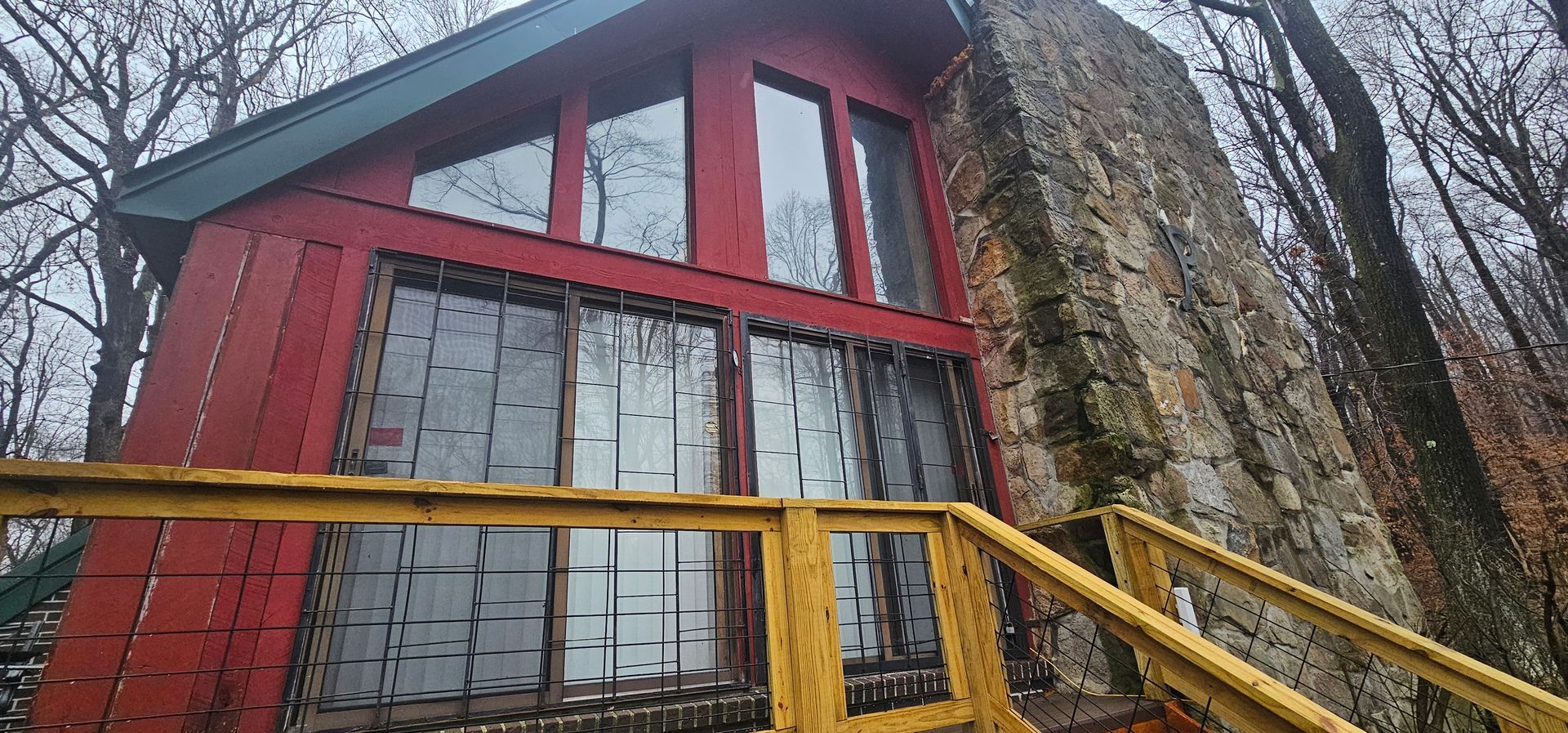 Red cabin with large windows and stone chimney, set in a wooded area with yellow railing and a green roof.
