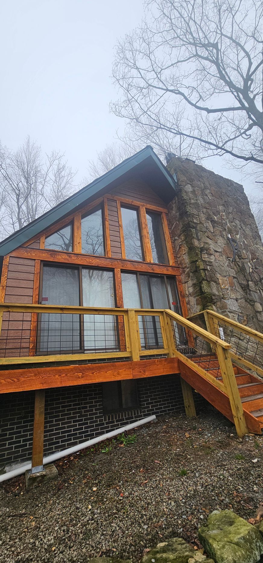 A-frame cabin with a large stone chimney, wooden deck, and cloudy sky.