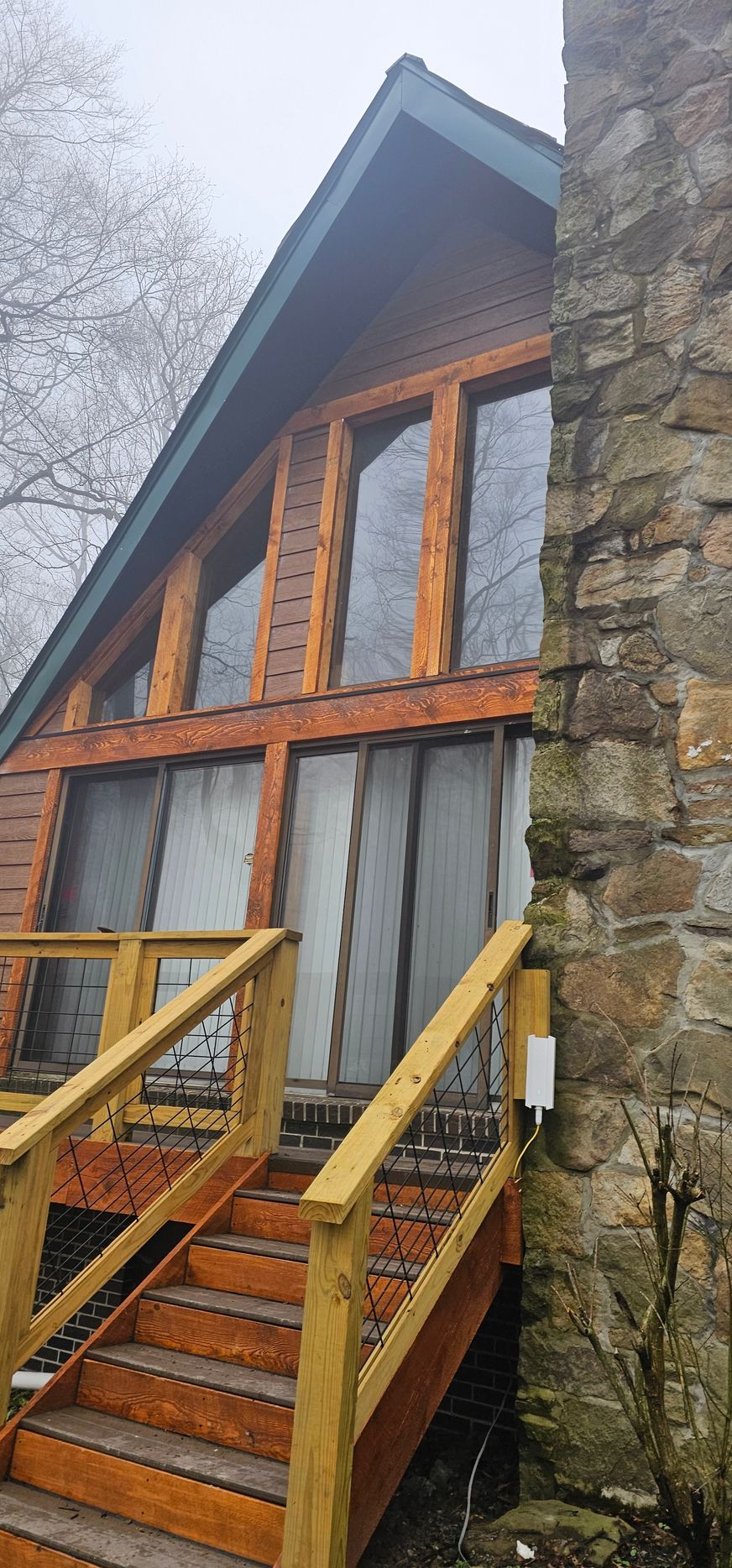 A-frame cabin with wooden deck and stone chimney. Brown wooden stairs lead up to the entrance with large windows.