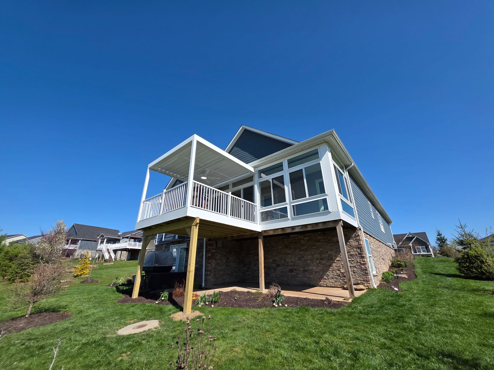 Back of house with deck and sunroom, blue sky overhead, grassy hillside.