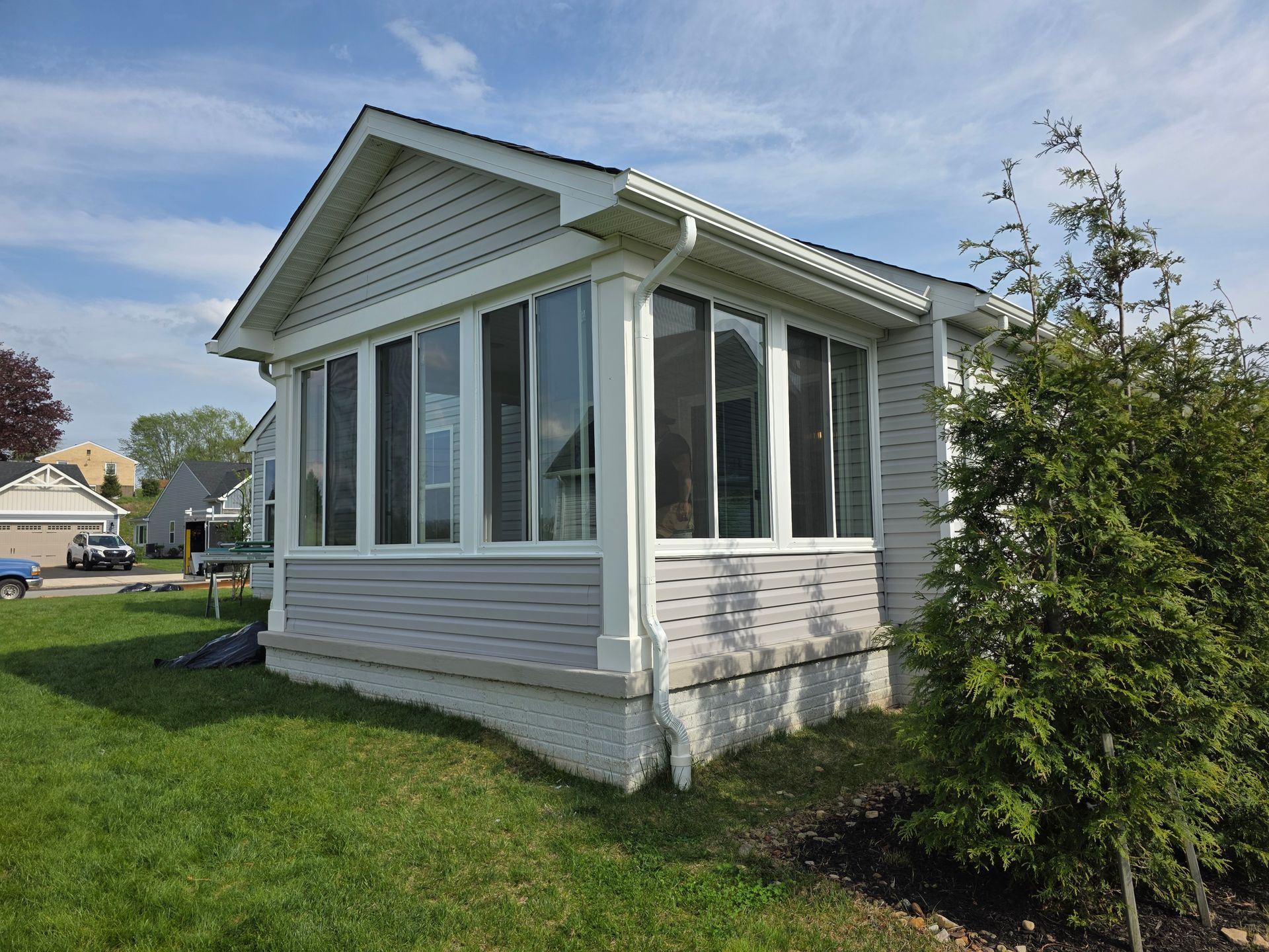 Sunroom with white trim, light gray siding, and many windows on a green lawn under a blue sky.