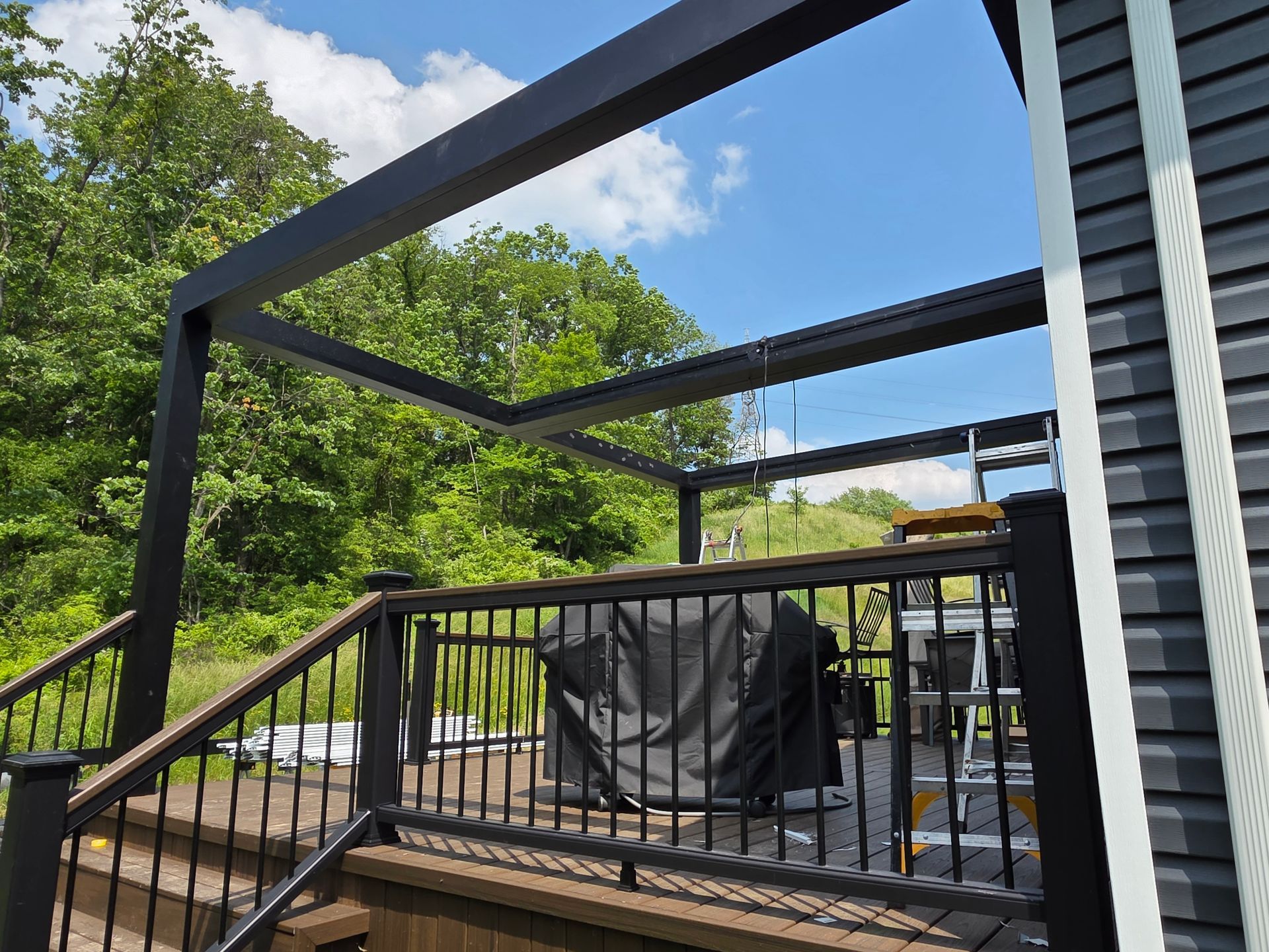 Black pergola over a deck with black railing and grill, against a backdrop of trees and blue sky.