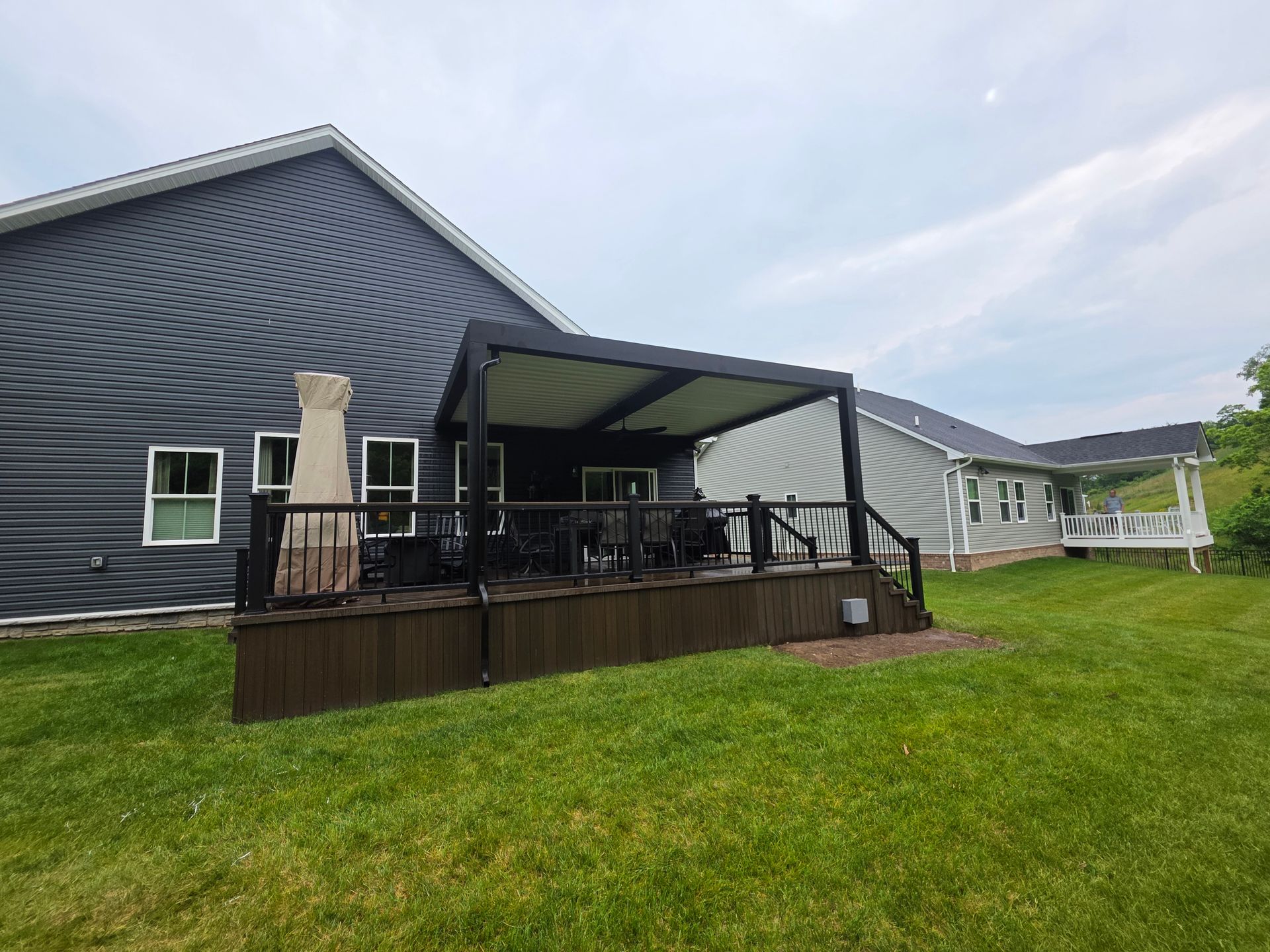 A backyard with a dark brown deck, black pergola, and gray siding on a house. Green grass in foreground.