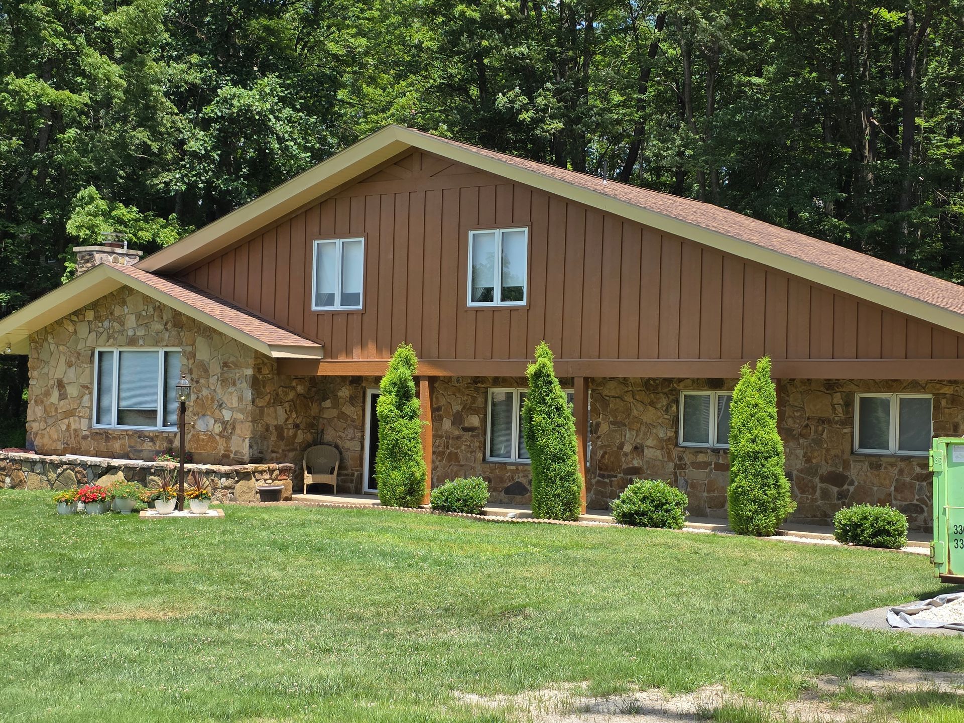 House with tan stone and brown siding, surrounded by trees and green lawn.
