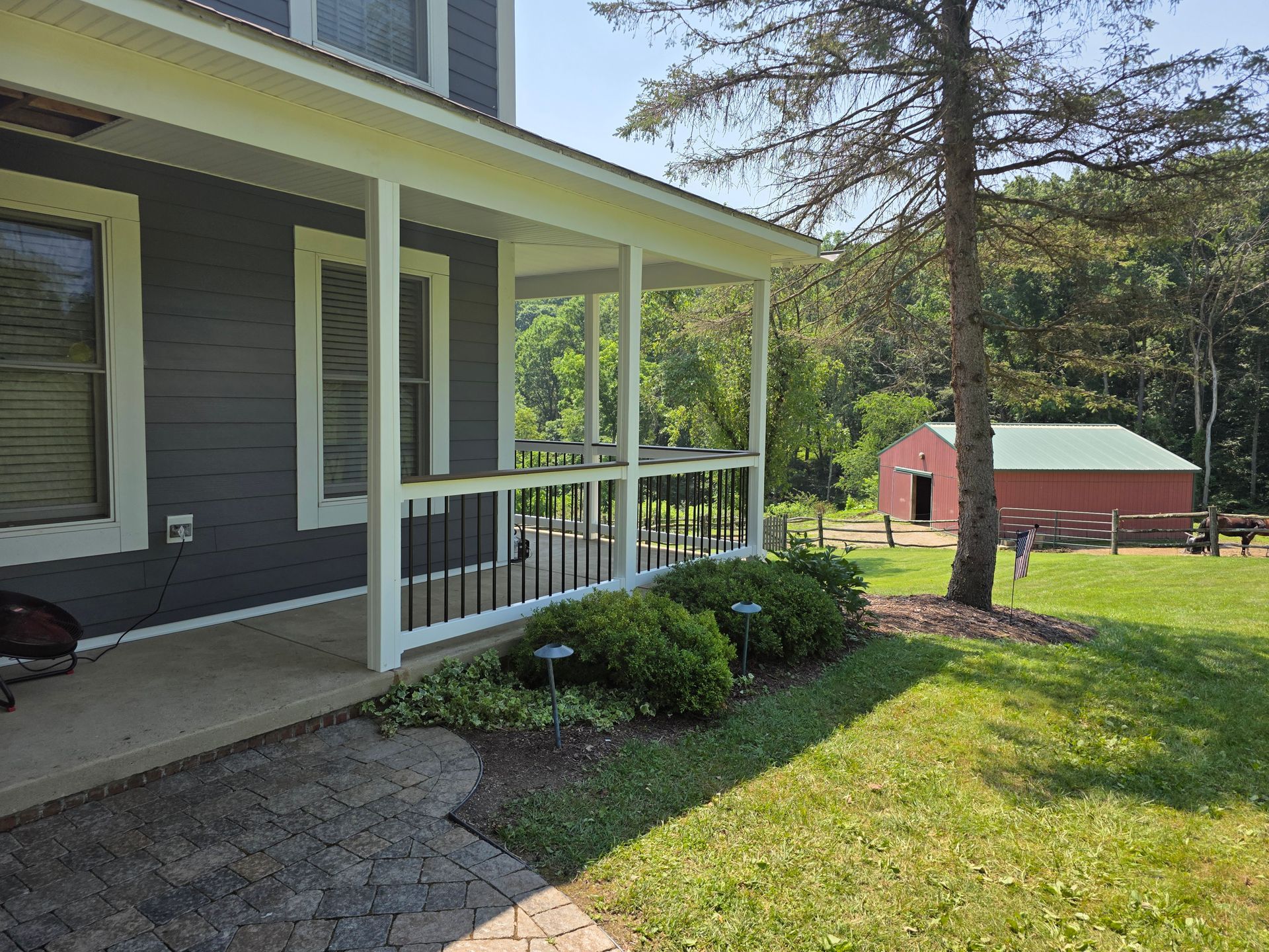 Gray house with a porch, a red barn in the background, and green trees on a sunny day.