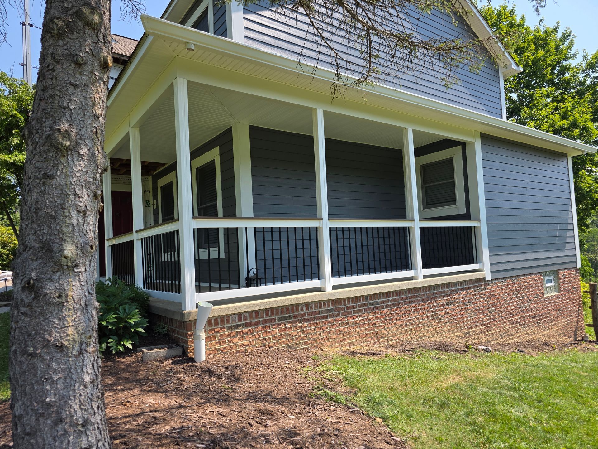 Covered porch of a two-story blue house with white trim and black railing. Tree in foreground.