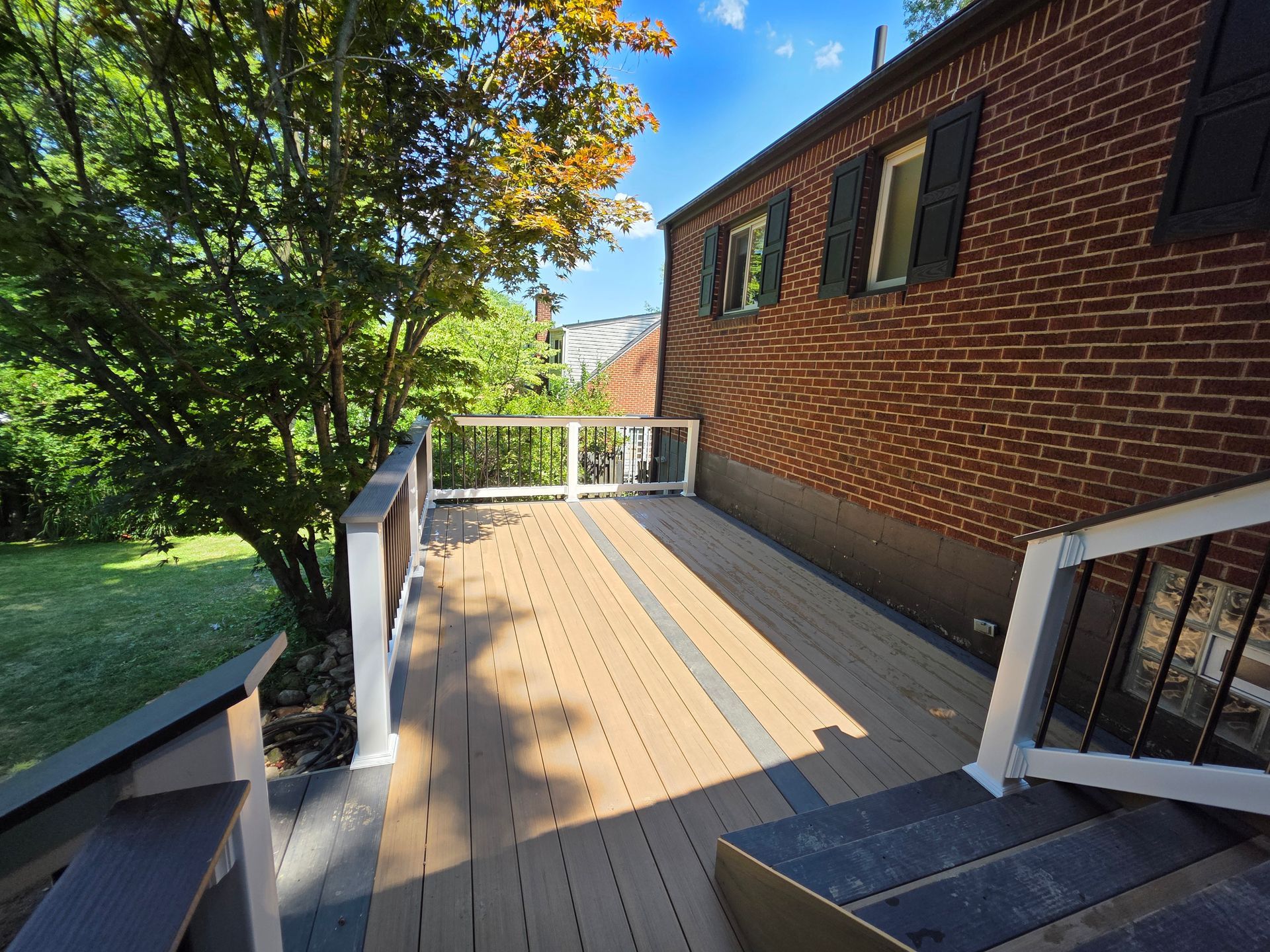 Wooden deck next to brick house with black shutters; steps lead down to a grassy yard.