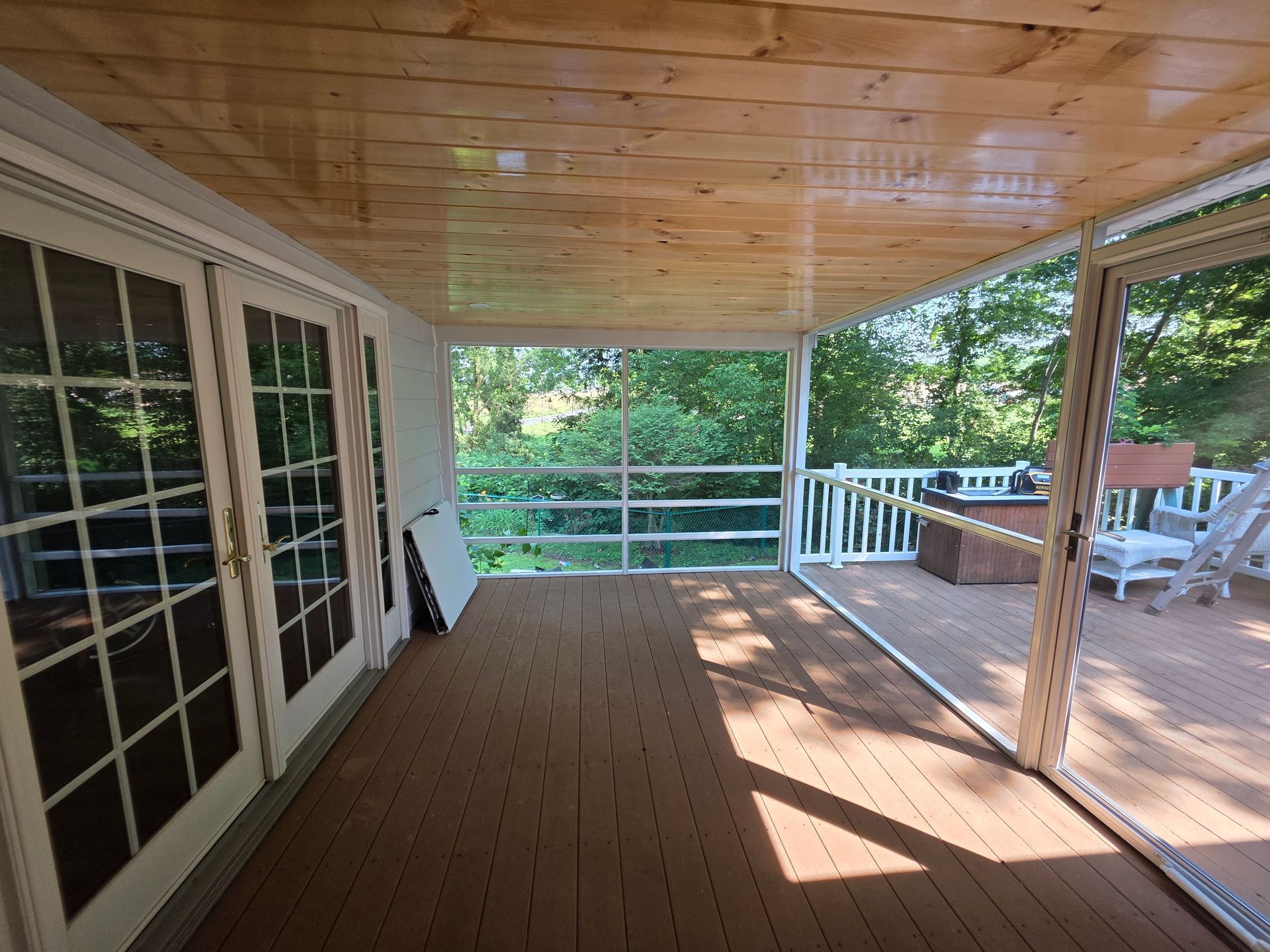 Covered porch with wood ceiling and flooring, white railing, and glass doors overlooking greenery.