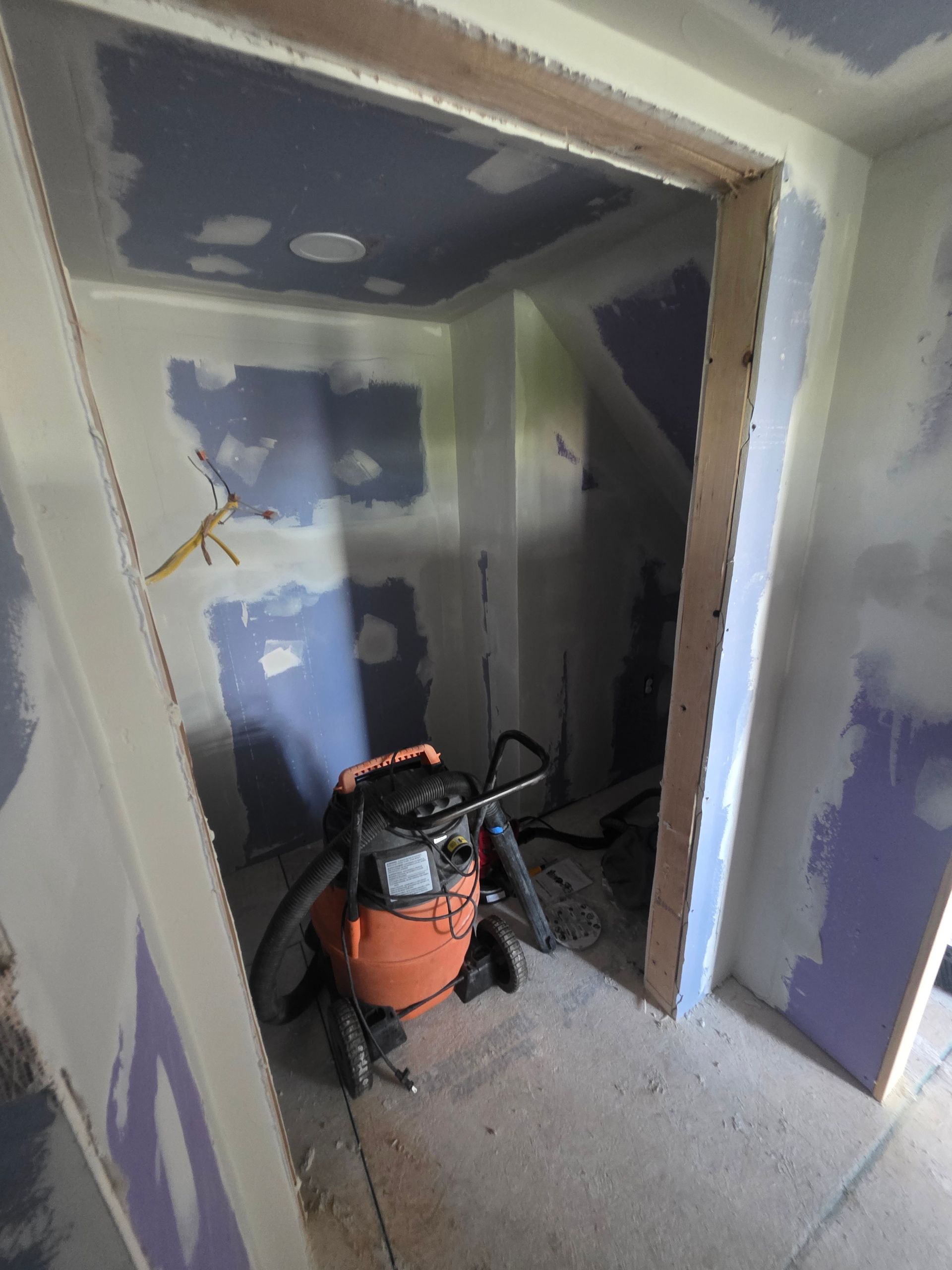 An orange shop vacuum sits inside a doorway of a room with unfinished drywall.