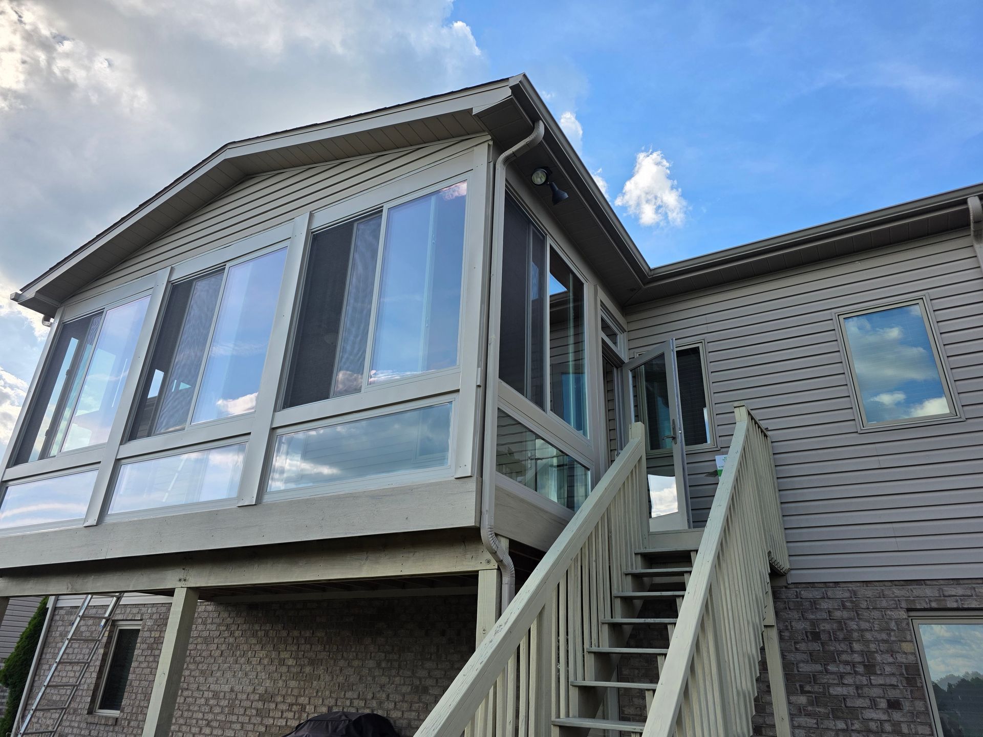 A sunroom with large windows and a staircase, attached to a house with siding, under a partly cloudy sky.