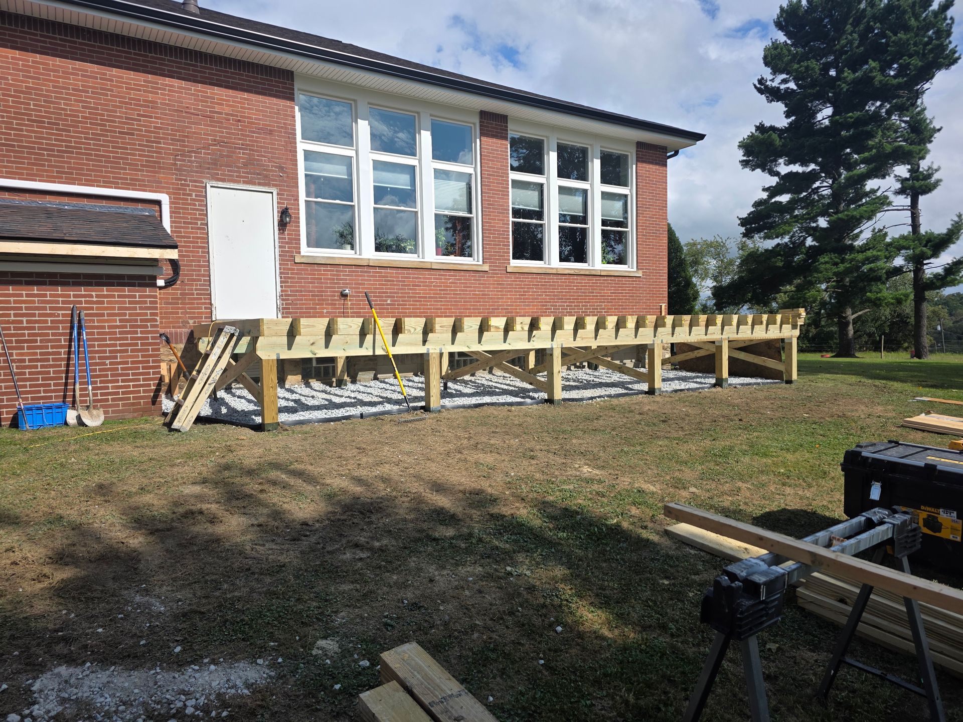 Deck construction in progress beside a brick building. Wooden frame assembled on gravel; unfinished.