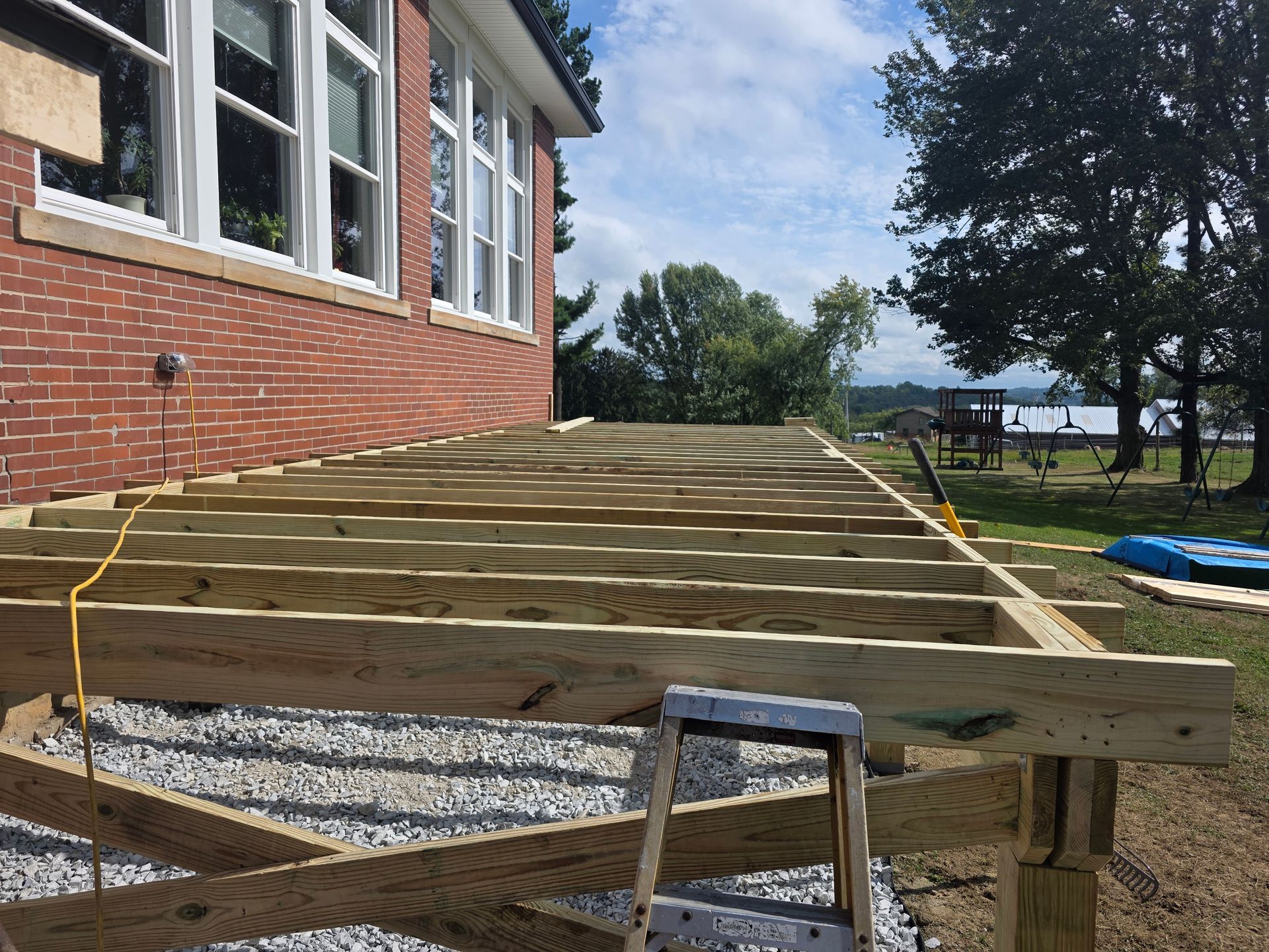 Deck construction in progress near a brick building. Wooden frame, gravel base, and ladder visible.