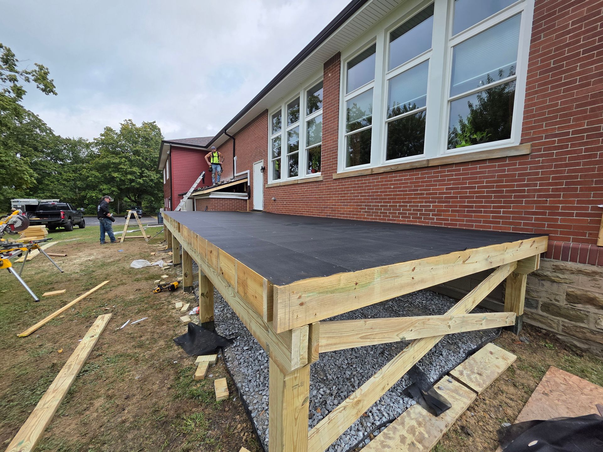 New wooden ramp construction outside a brick building. Workers are present, and gravel base is visible.