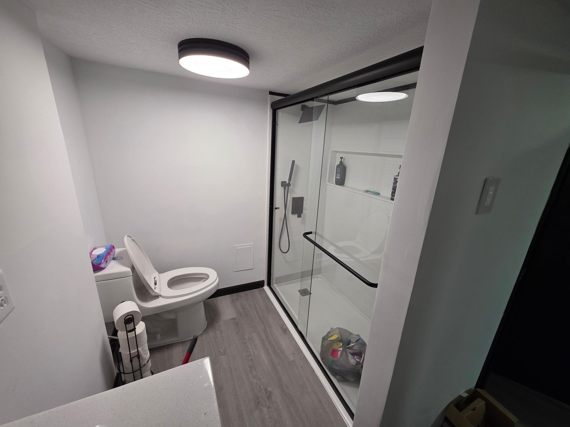 Bathroom with a toilet, shower, and a black-framed glass door. Gray floor, white walls and a light fixture.