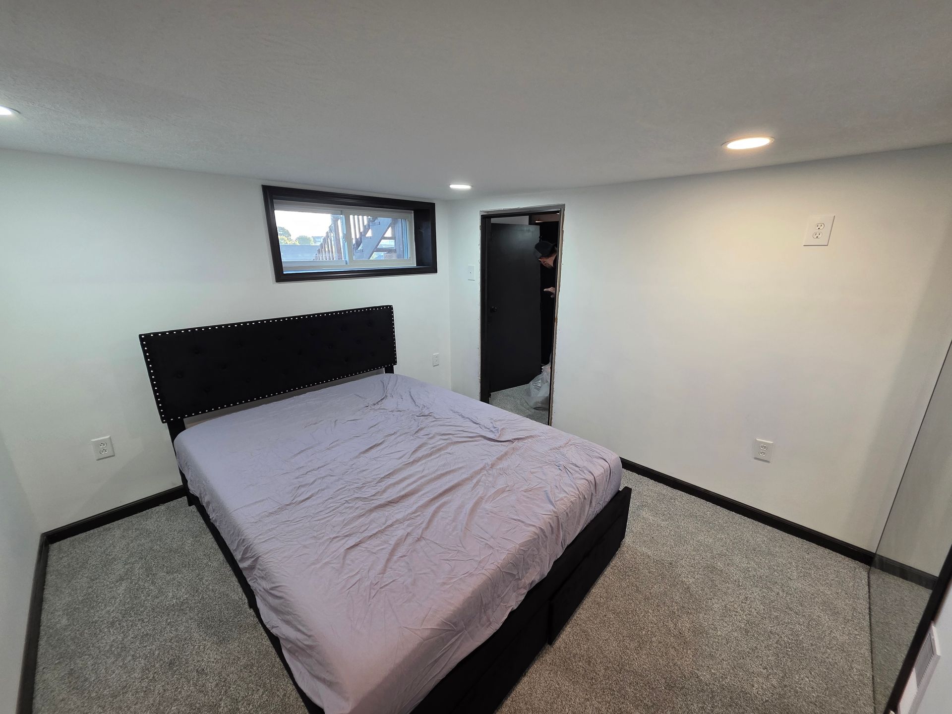 Bedroom with bed, small window, black door, and gray carpet.