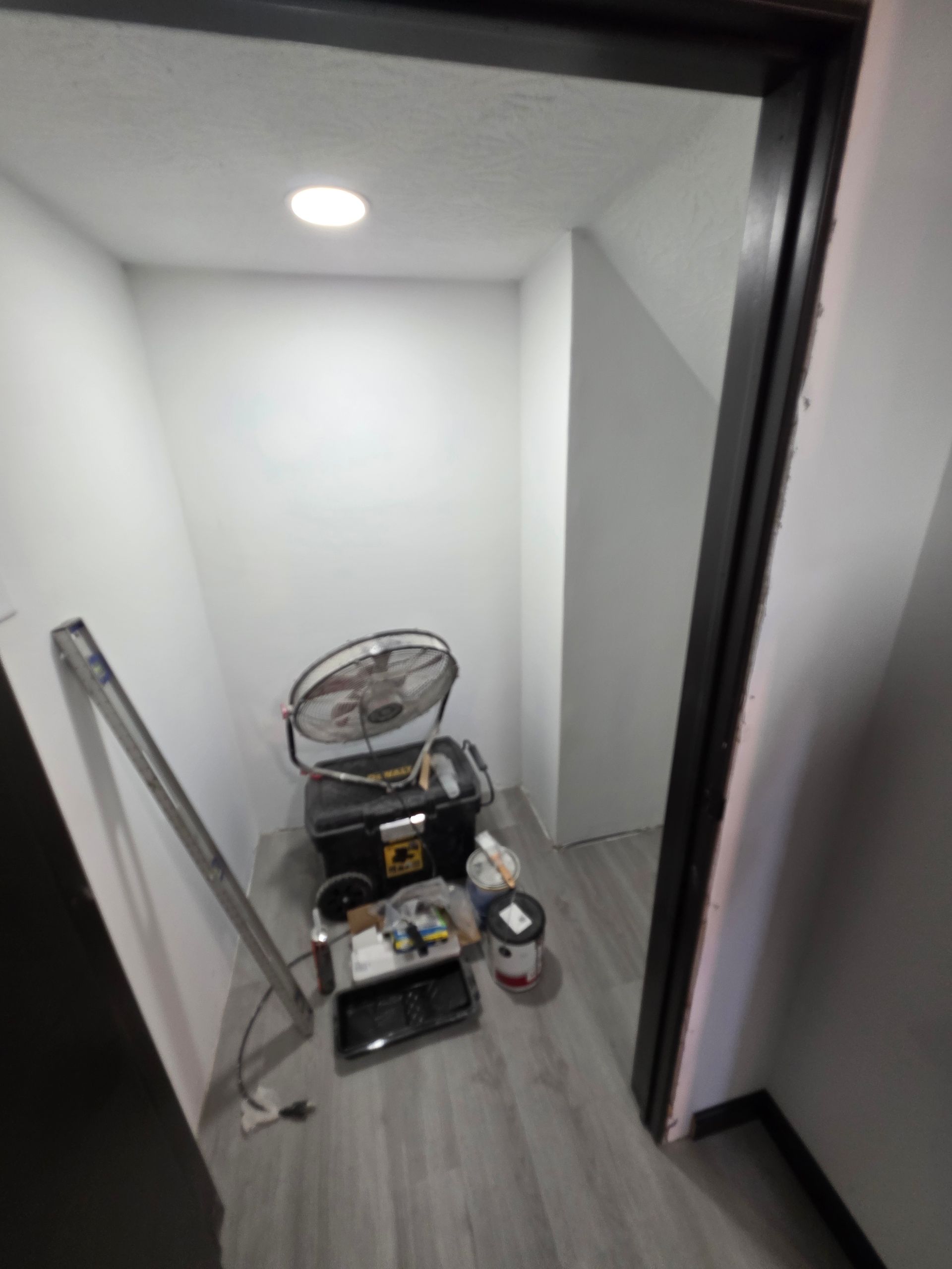 Small closet with tools, fan, and paint supplies on the floor. Gray flooring and white walls.