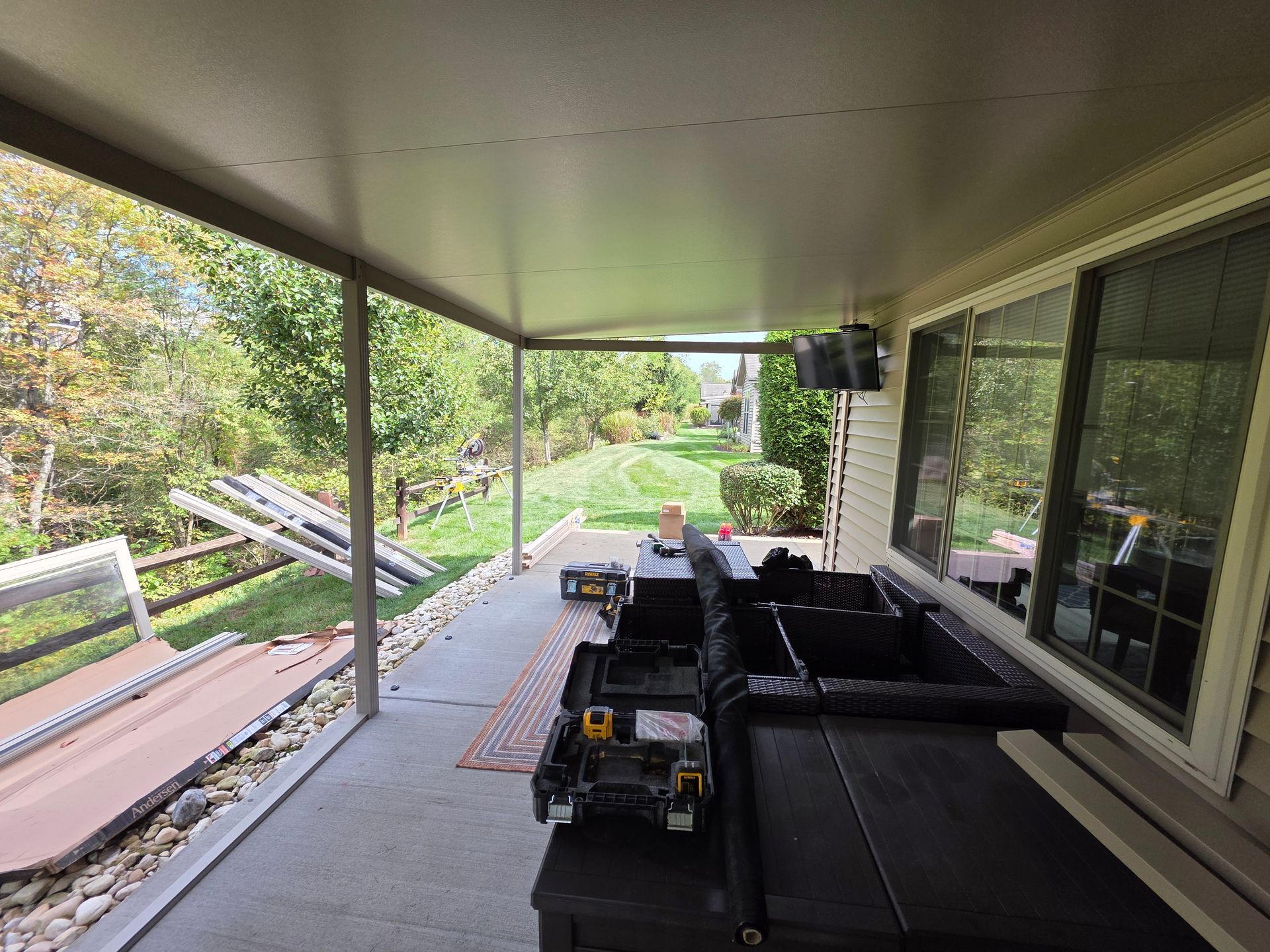 Covered patio with a black couch, tools, and a TV; a backyard with grass and trees.
