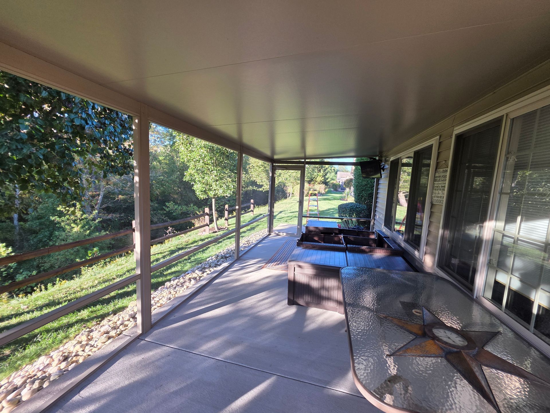 Covered patio with a view of trees, a stone and wooden fence, and a glass table.
