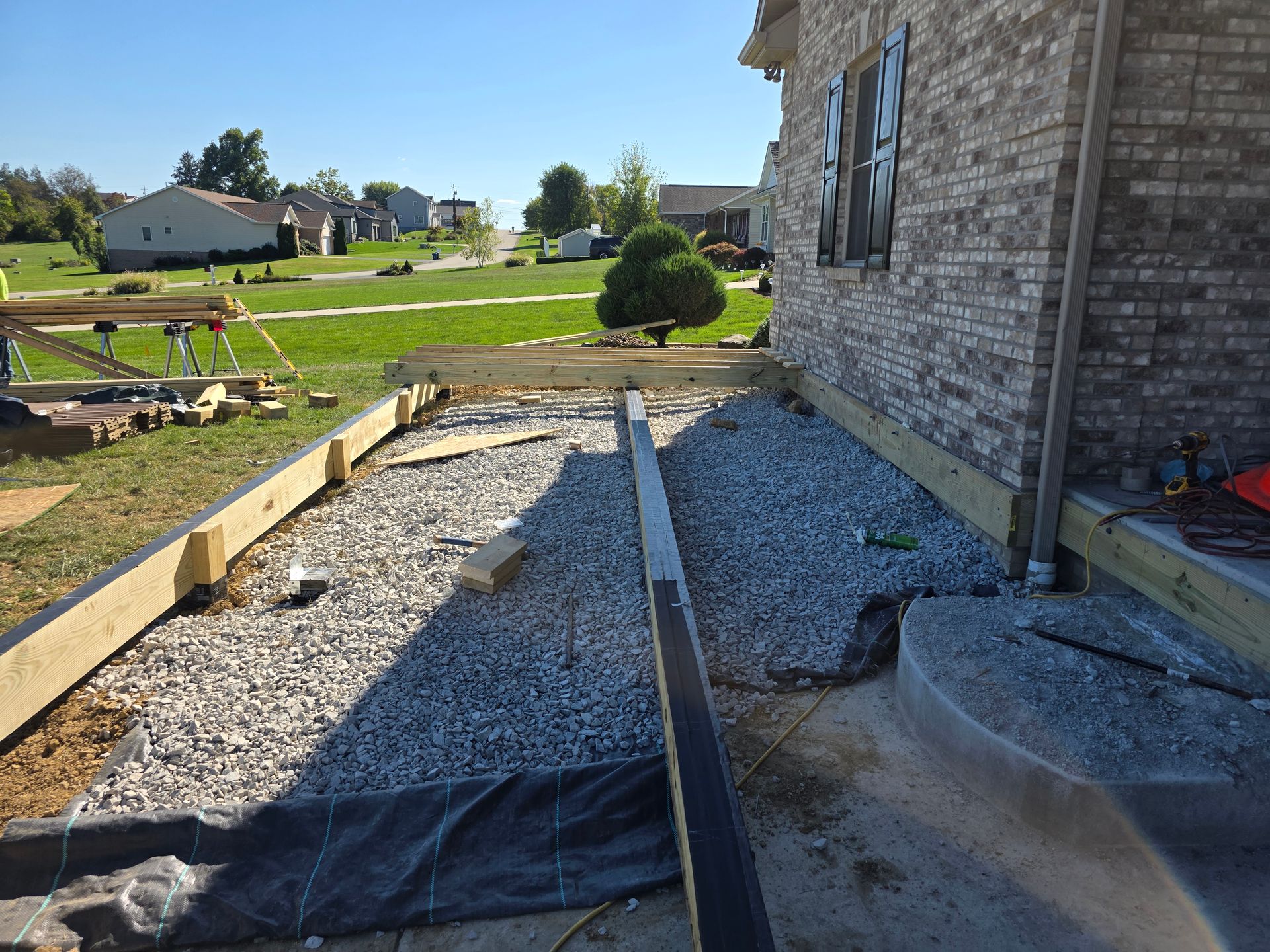 Construction site with gravel, wooden beams, and a house with brick siding.