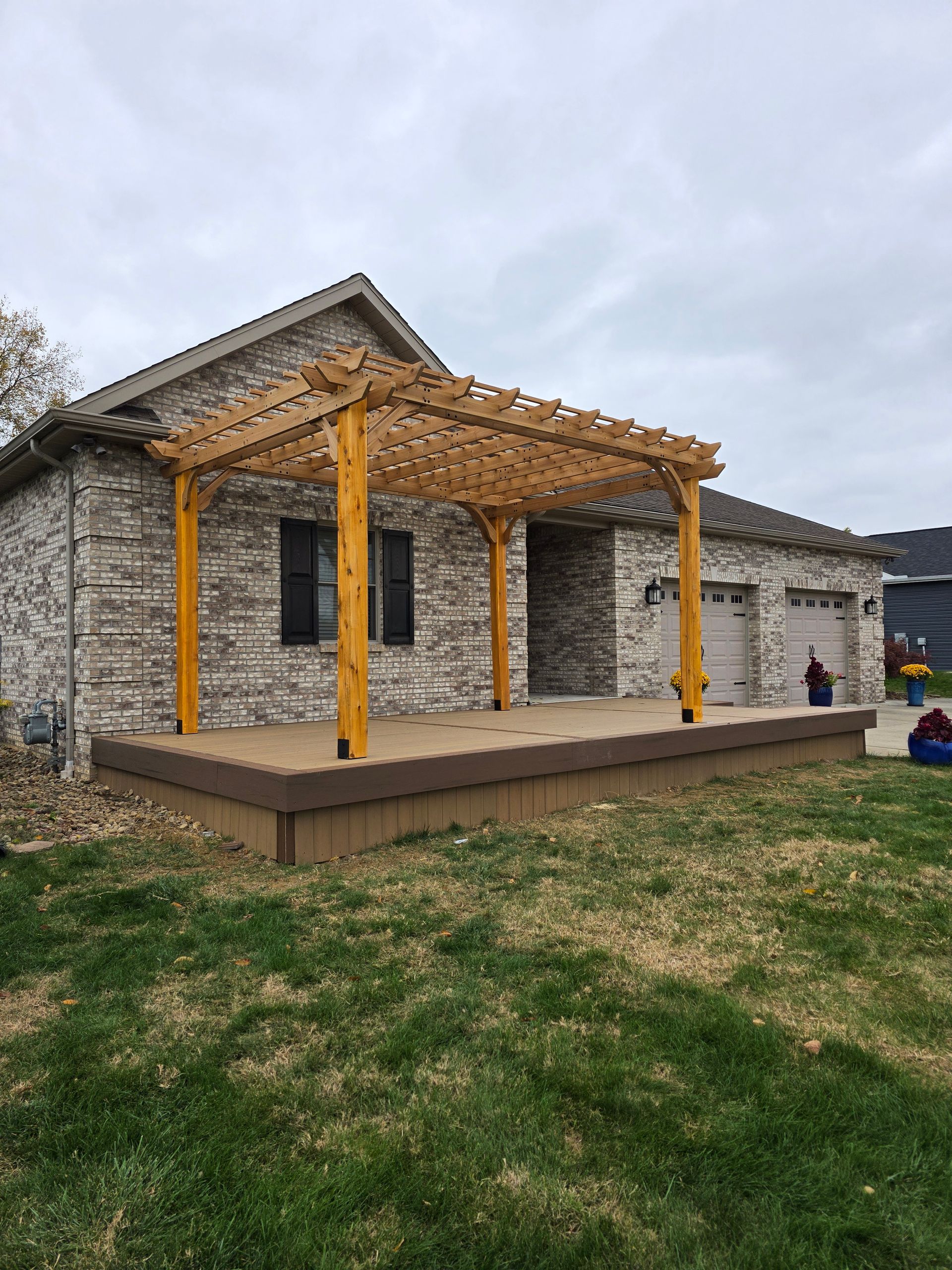 Wooden pergola over a deck in front of a brick house on a cloudy day.