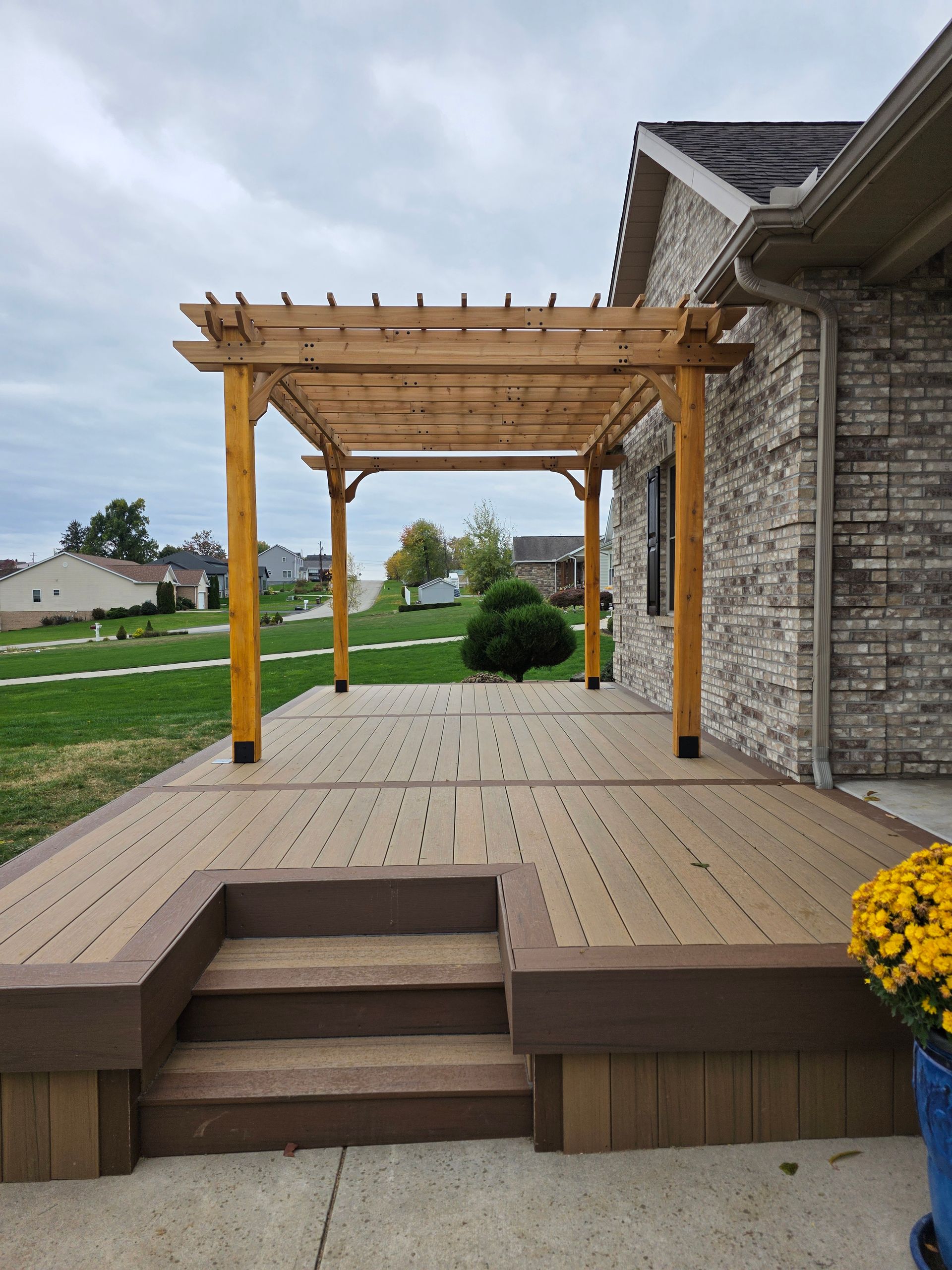 Wooden pergola over a composite deck with steps. A brick house is to the right, and grass and trees are in the distance.