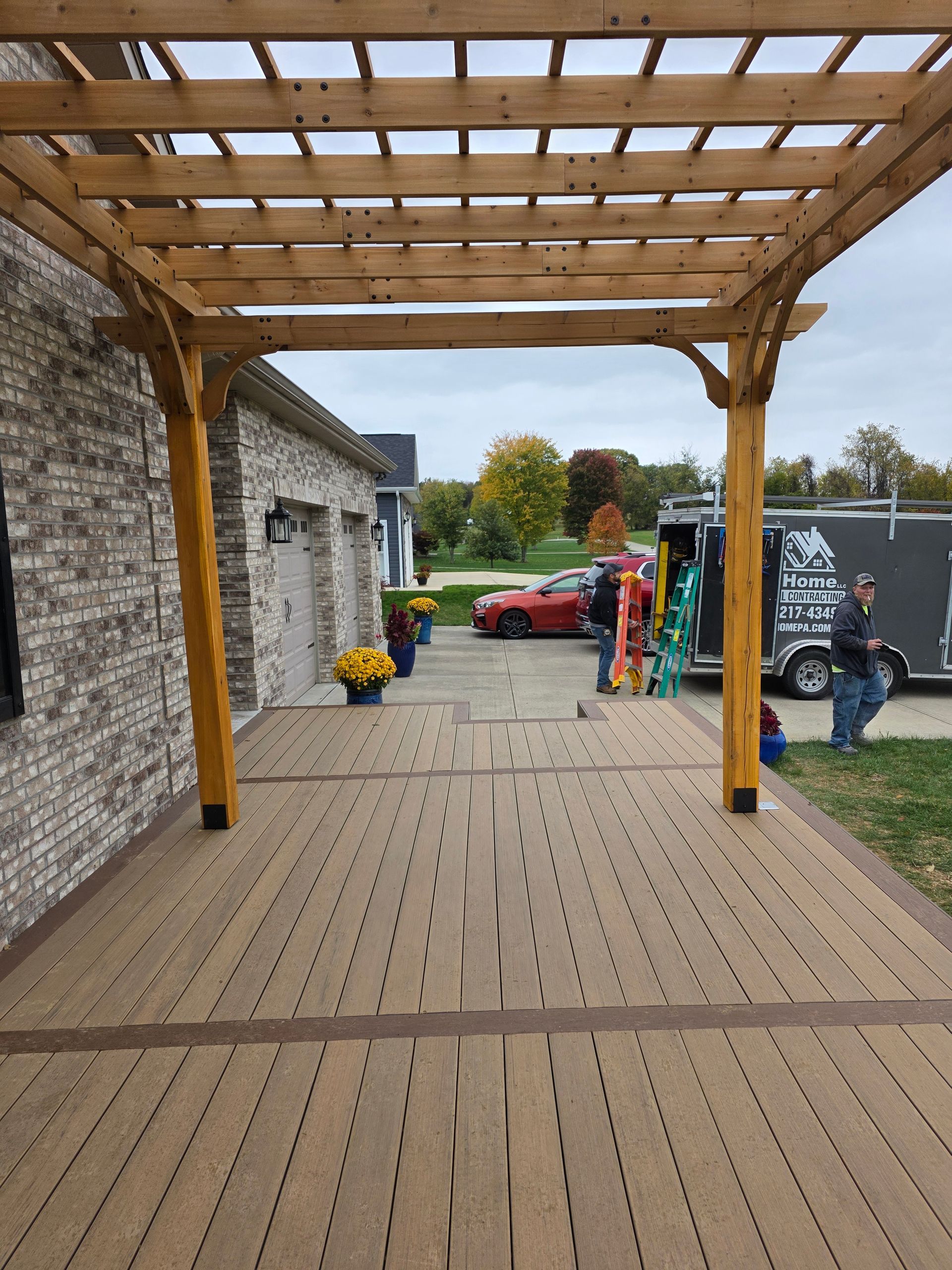 Wooden pergola over a composite deck. A brick building with garage doors is on the left; a red car is in the background.