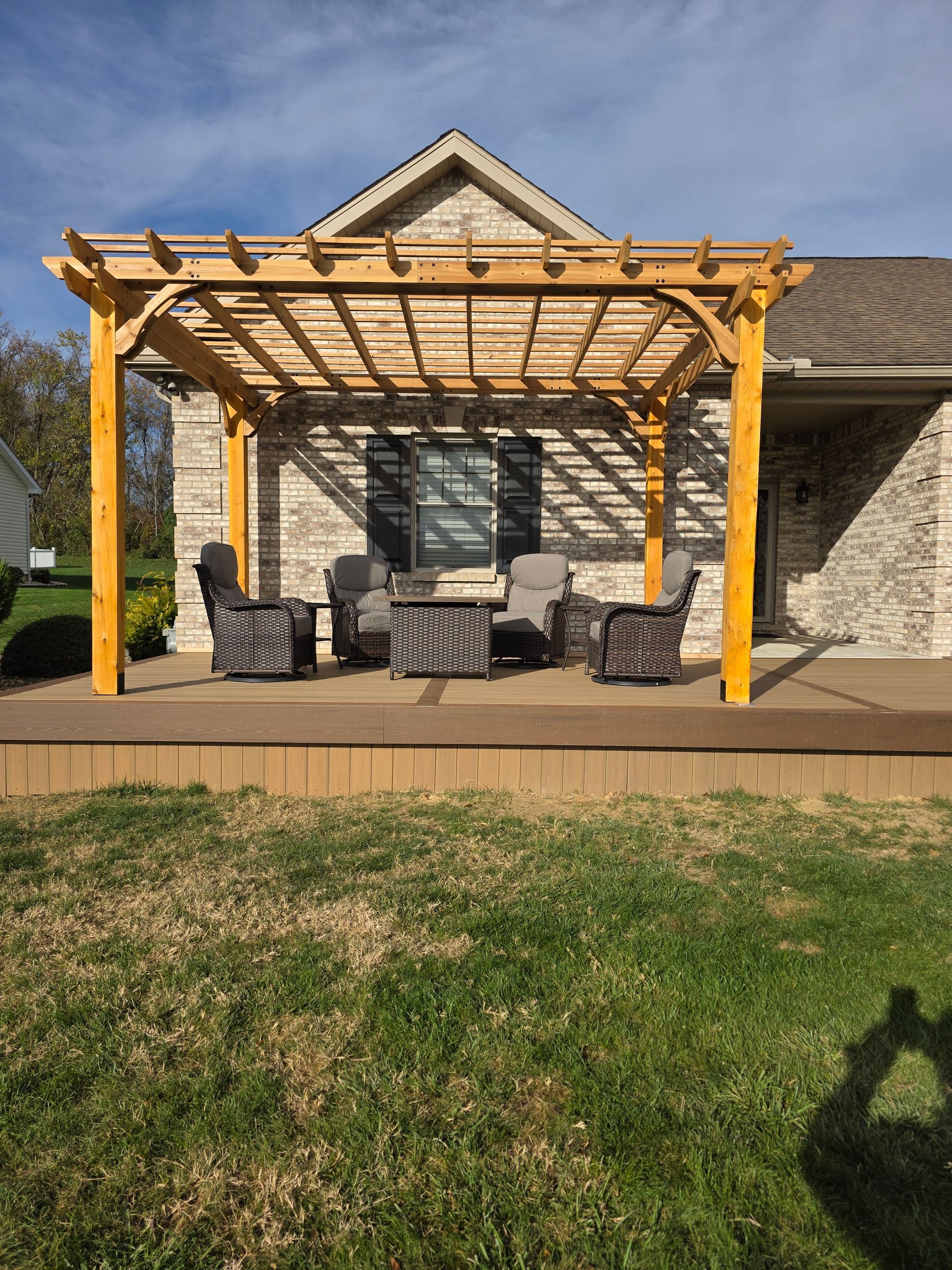 Wooden pergola over outdoor seating on a deck, in front of a brick house.