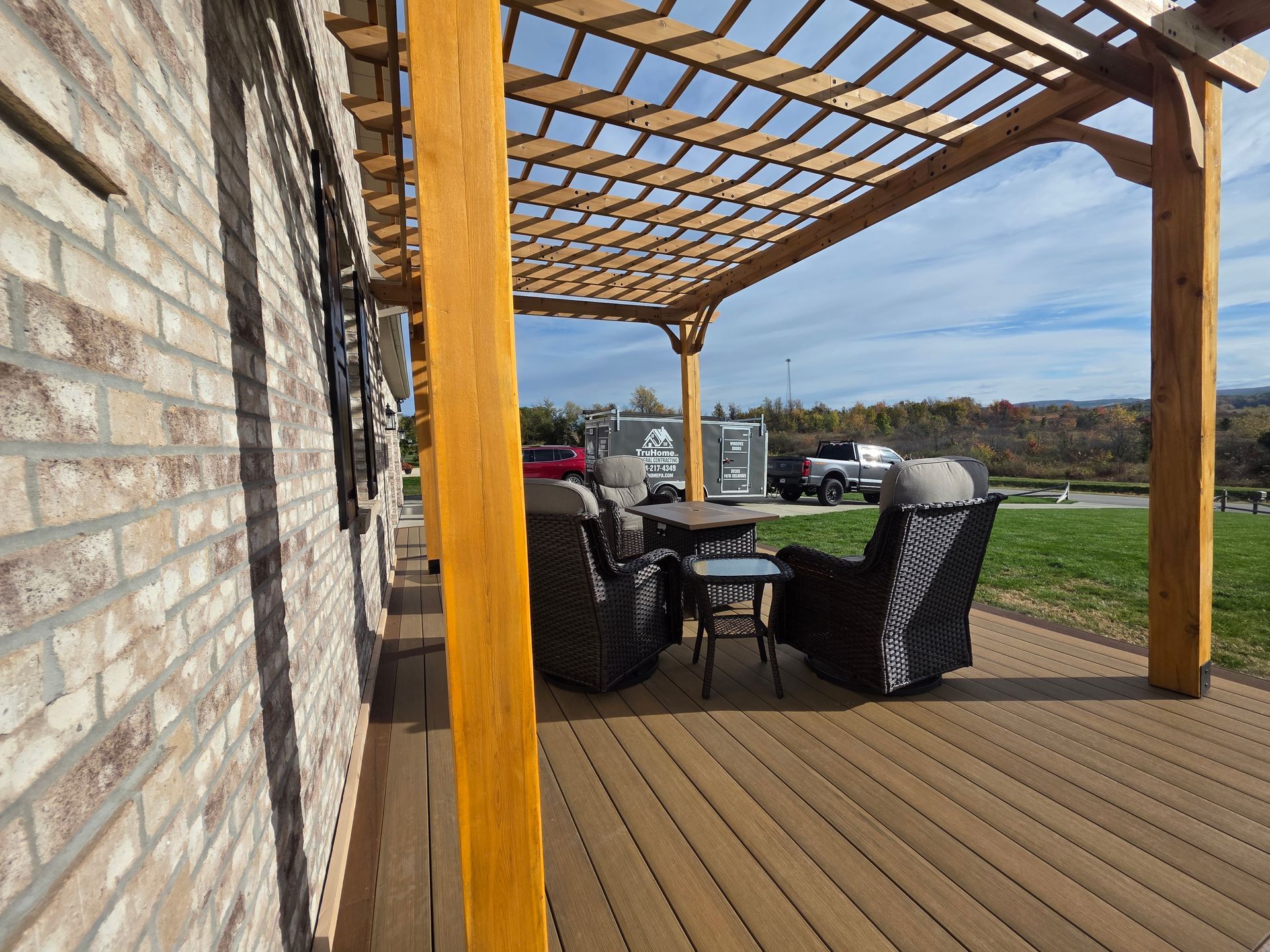 Wooden pergola over a deck with two chairs, table, brick wall, and a view of a field.
