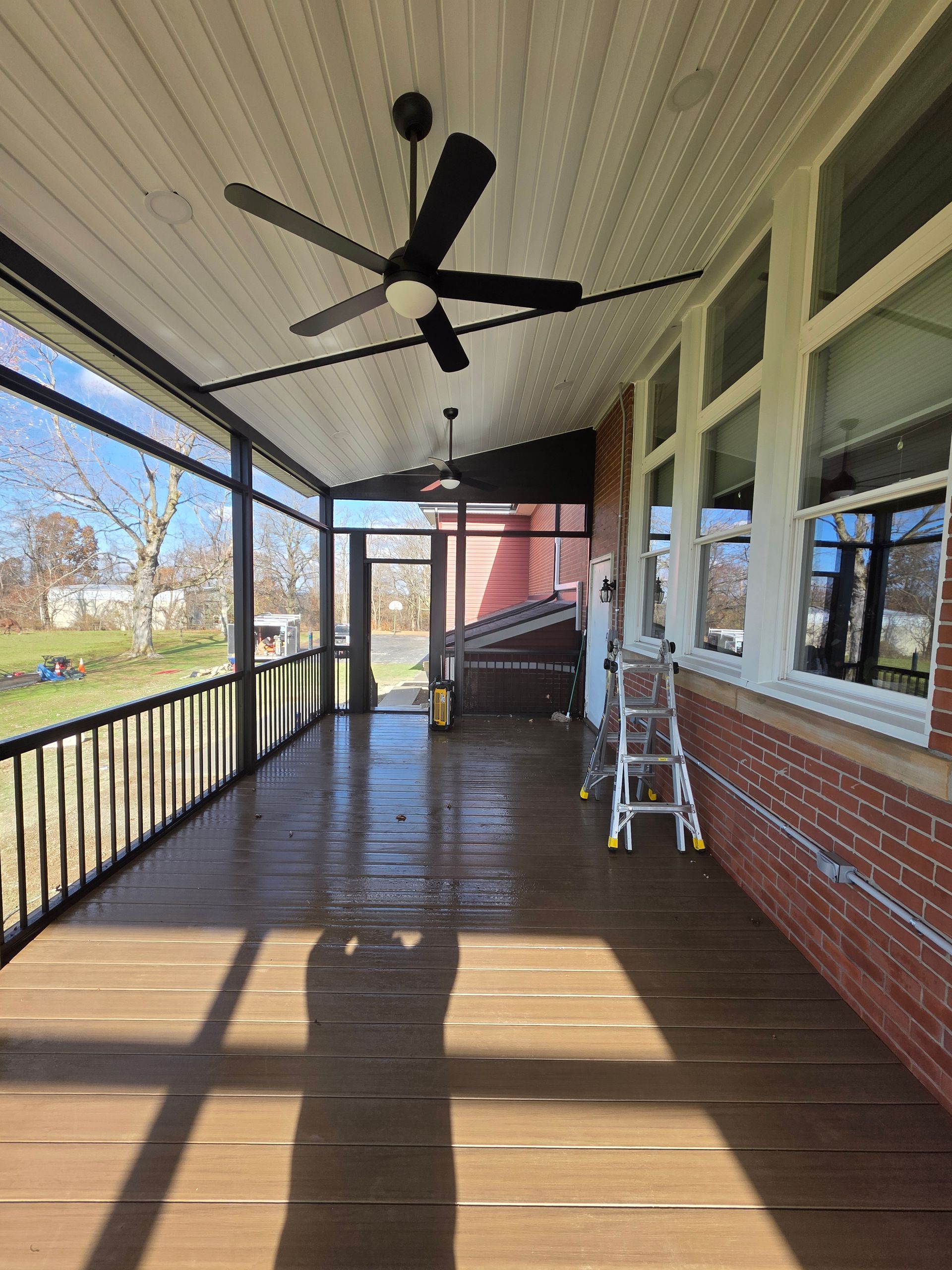 Covered porch with wood flooring, screen panels, and two ceiling fans. A red brick wall is on the right.