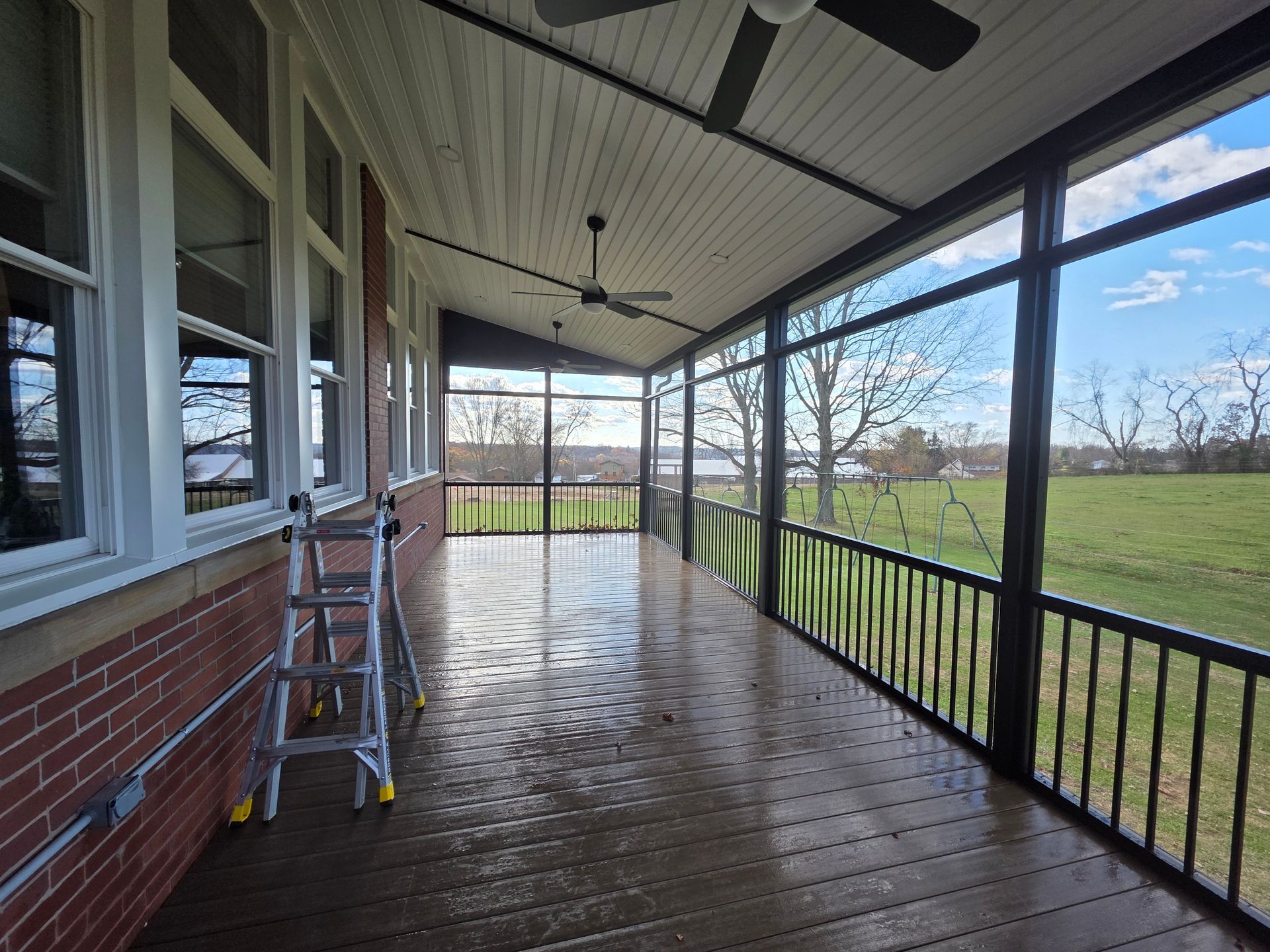 Covered porch with wet wood flooring, screened sides, brick wall, white ceiling.  Ladder present.